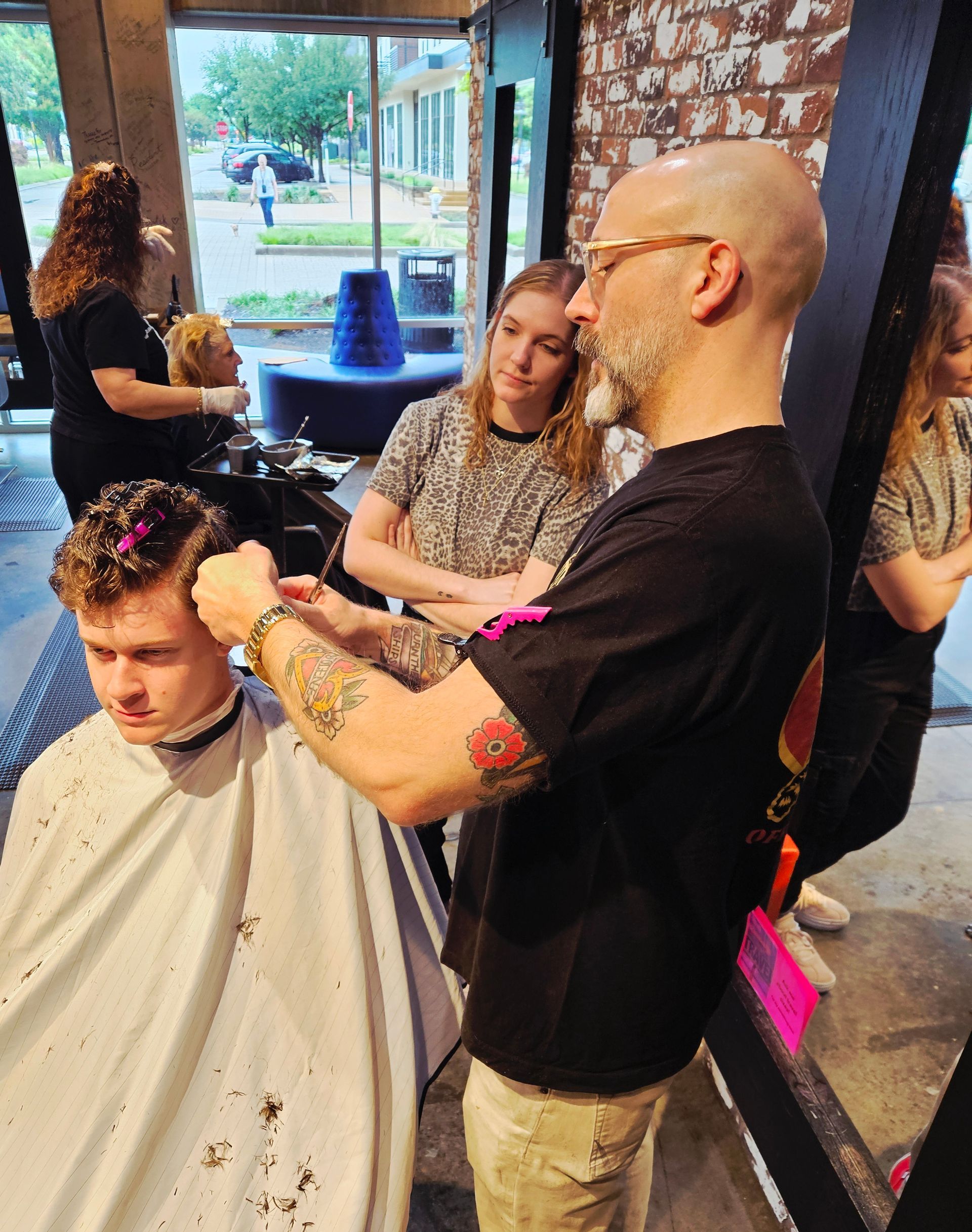 Hair stylist cutting client's hair in a salon, another person watches. Light interior with windows.