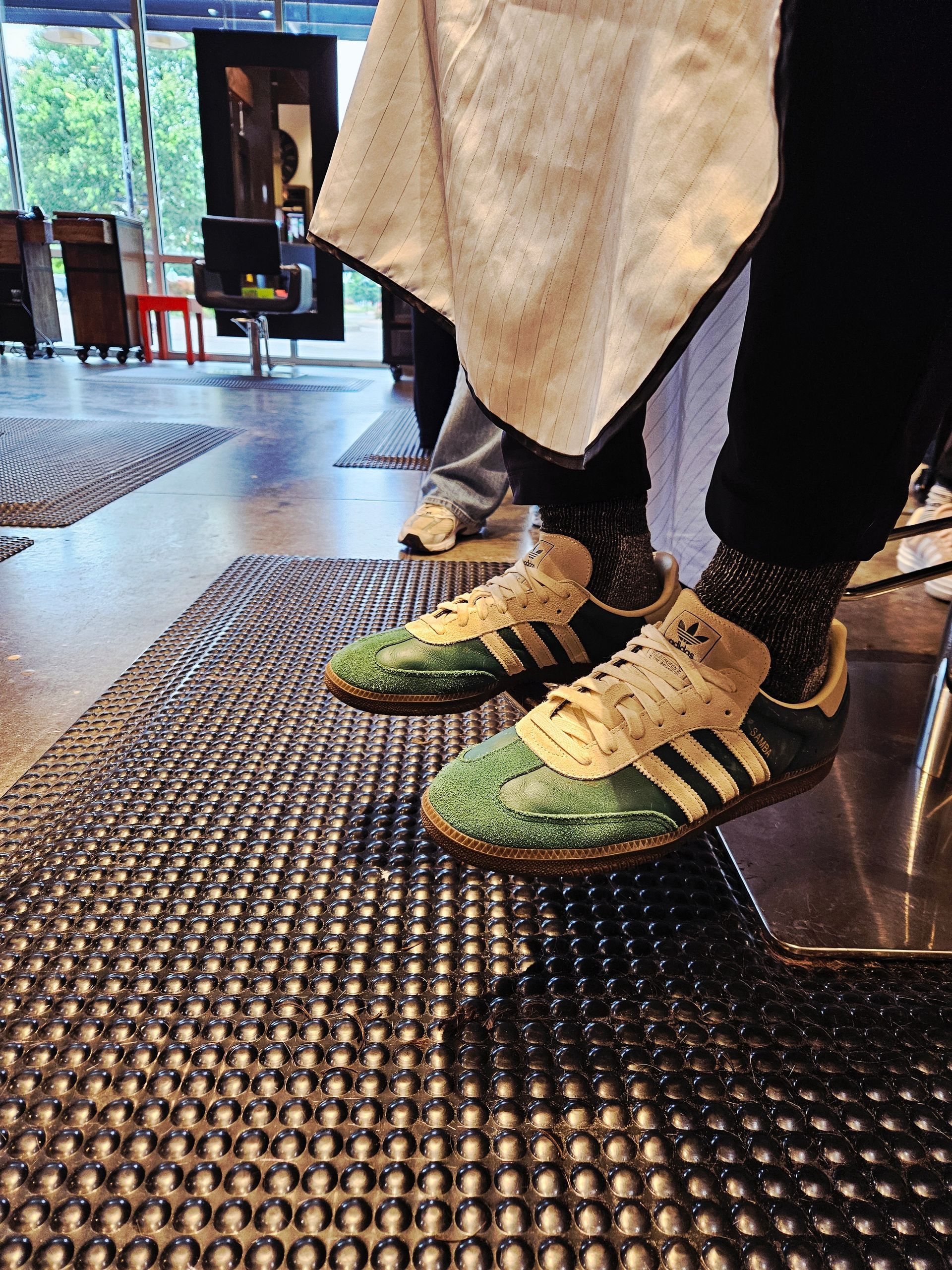 Person wearing green and white Adidas sneakers stands on a textured mat in a barber shop.