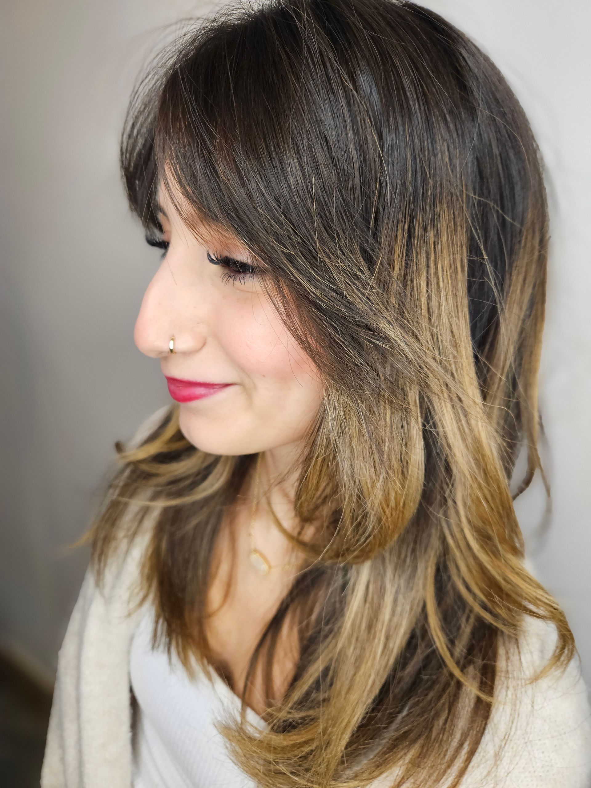 Woman with brown hair, face framed by layers, light brown and blonde highlights, wearing nose ring, smiling.