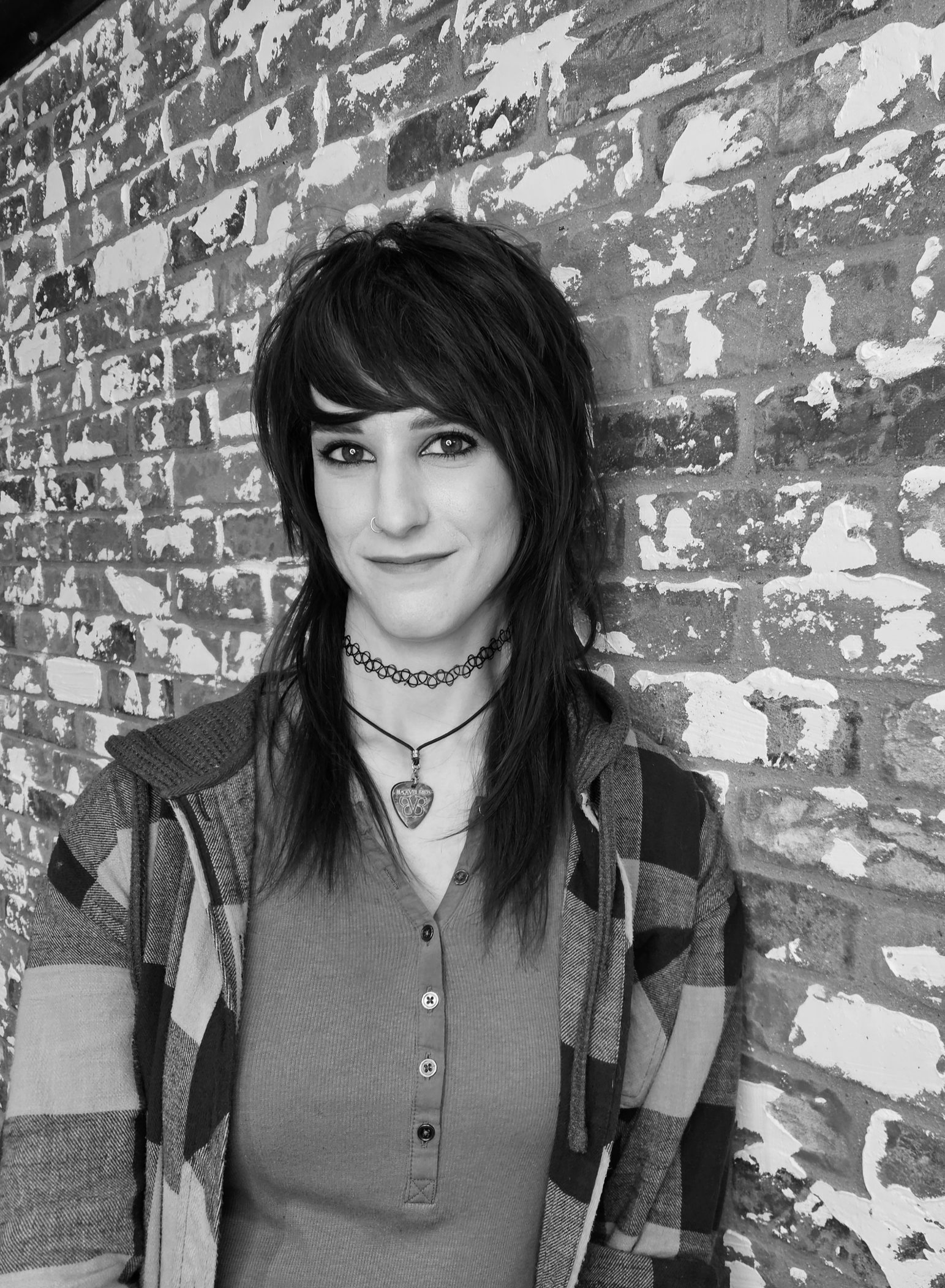 A person with dark hair and a layered, choppy haircut stands against a textured brick wall, wearing a layered outfit.