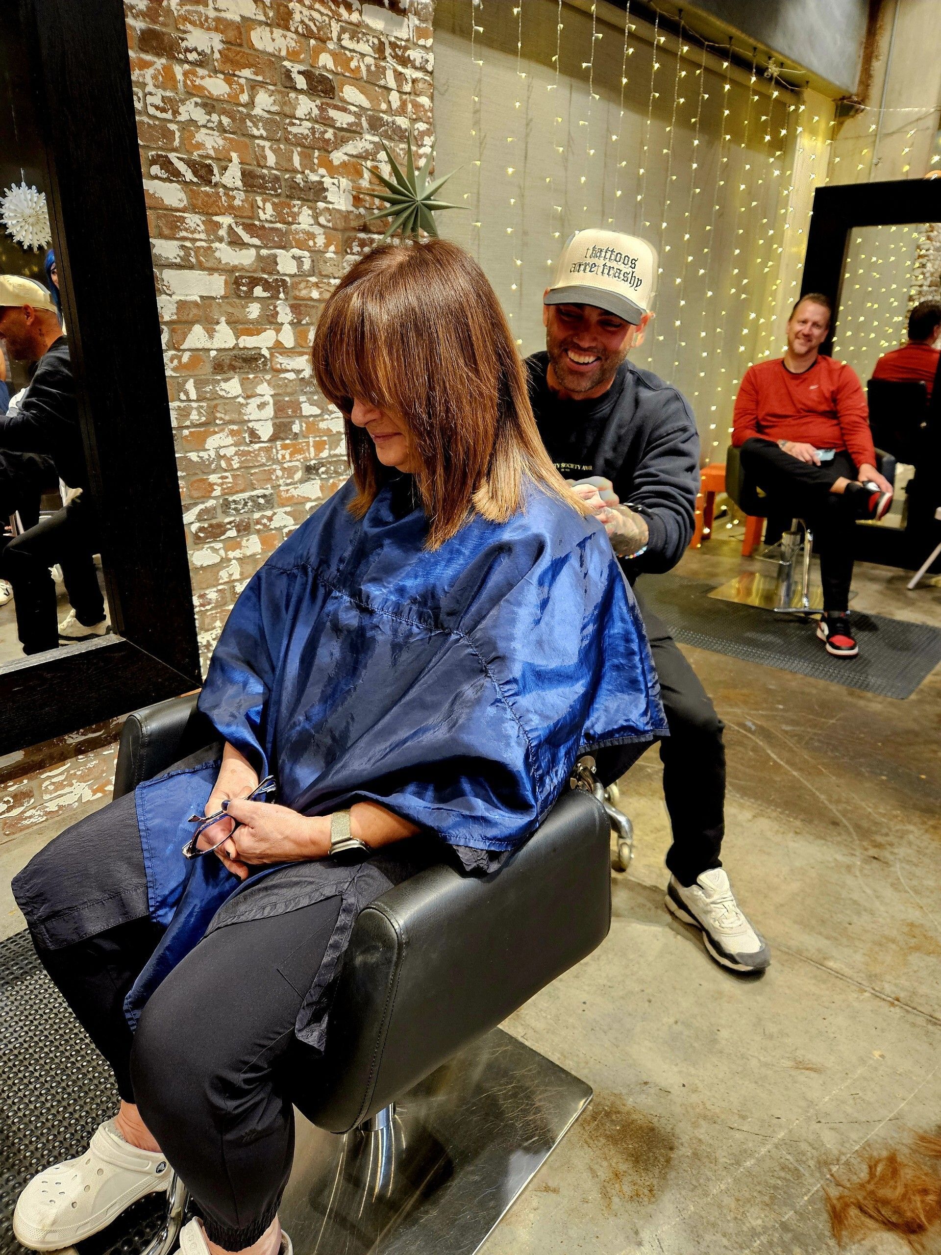 Hairdresser cutting a client's hair in a salon. The client is wearing a cape. Other person smiles in the background.