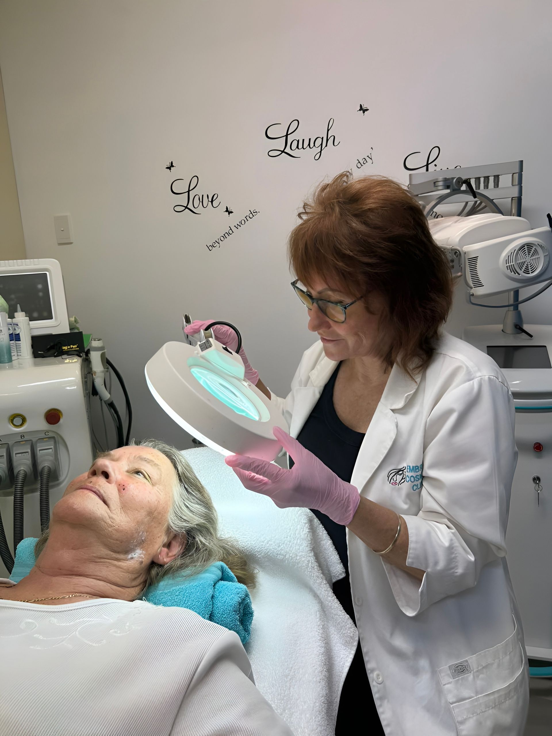 Woman in Lab Coat Examines Patient's Face With Magnifying Lamp — Embrace Cosmetic Clinic in Bundaberg North, QLD