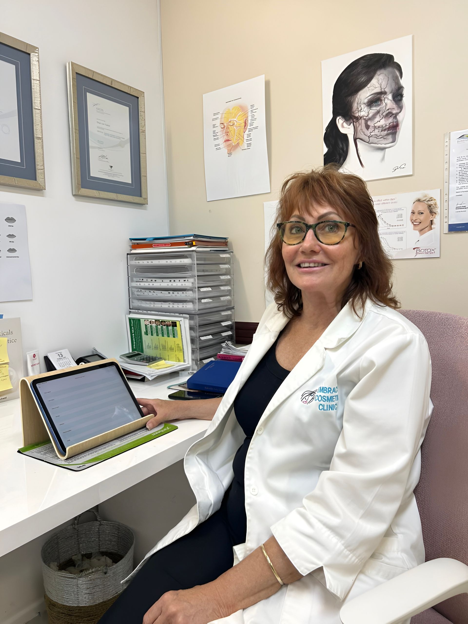 Woman in White Coat Smiles, Seated at a Desk — Embrace Cosmetic Clinic in Bundaberg North, QLD