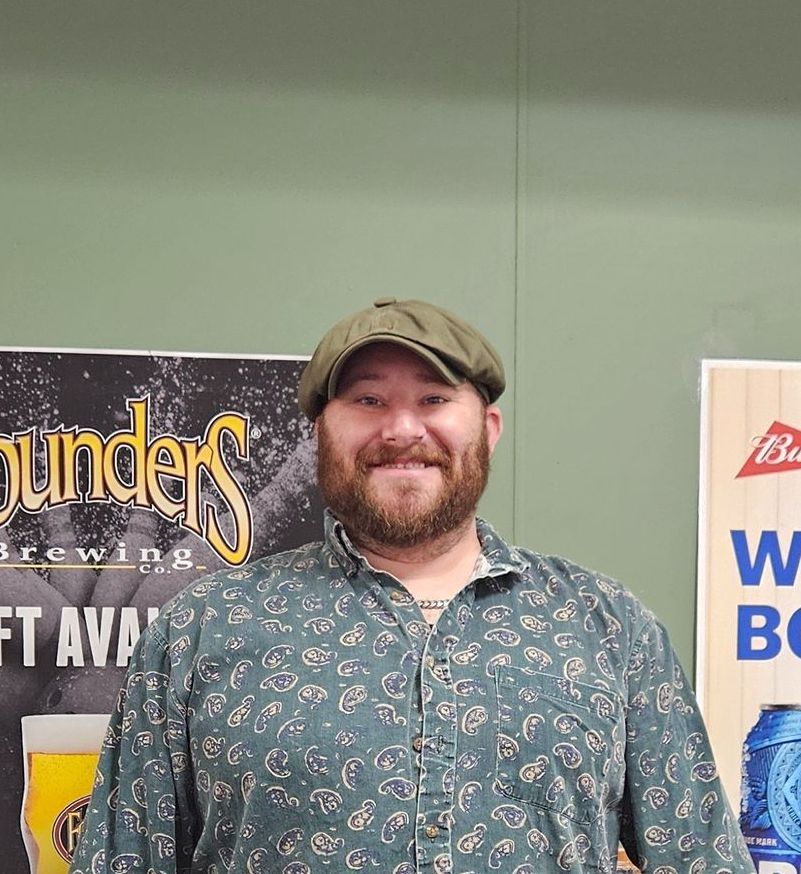 Man with beard and cap smiles. Standing in front of beer advertisements.