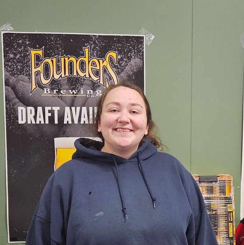 Woman smiles in front of a Founders Brewing poster. She wears a blue hooded sweatshirt.