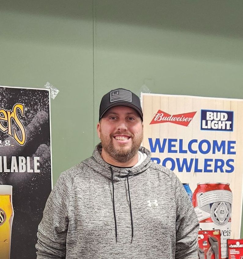 Man in a gray hoodie and cap smiles near a Bud Light sign that says 