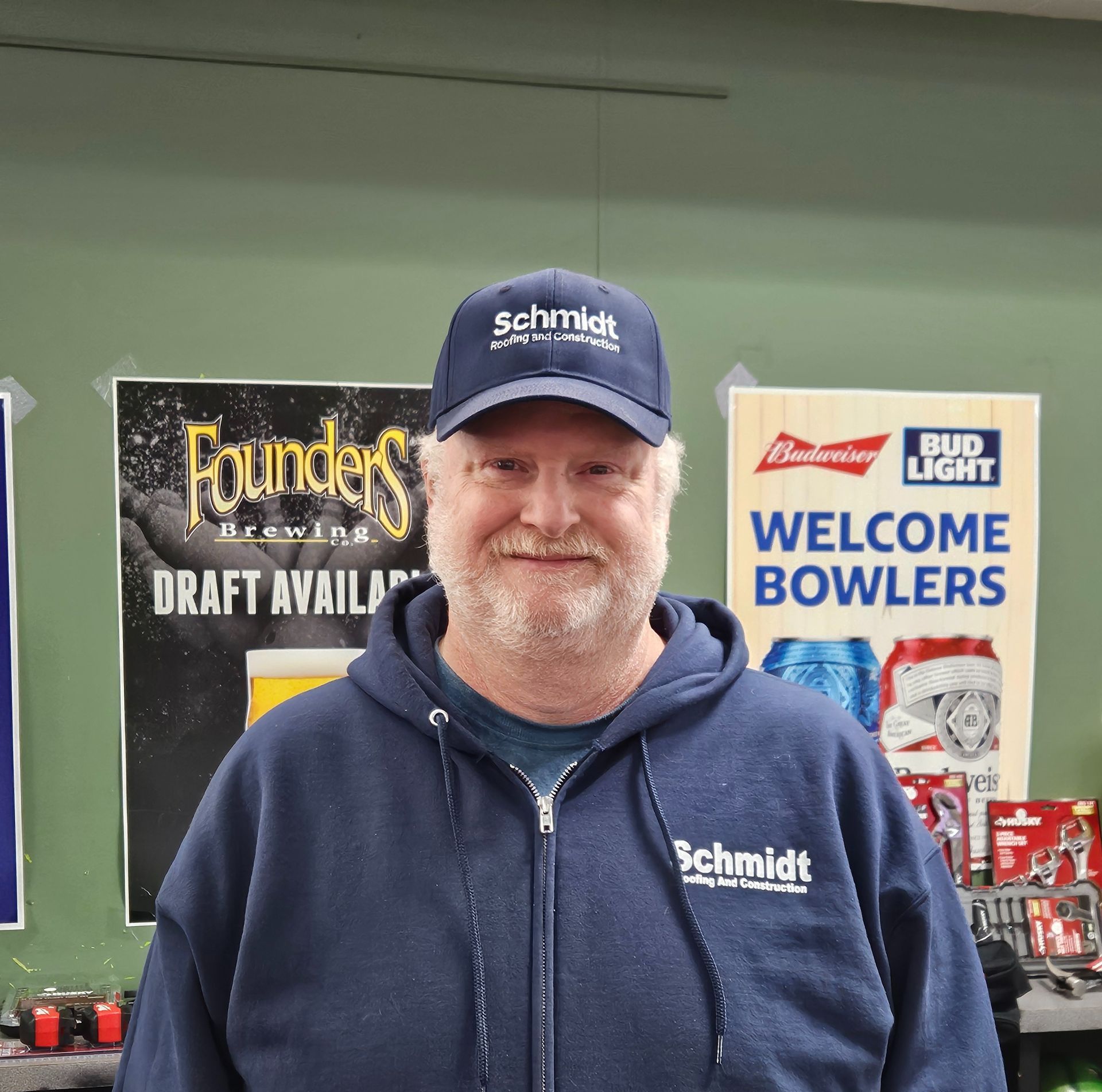 Man in a Schmidt logo cap and sweatshirt smiles in front of brewery and bowling signs.