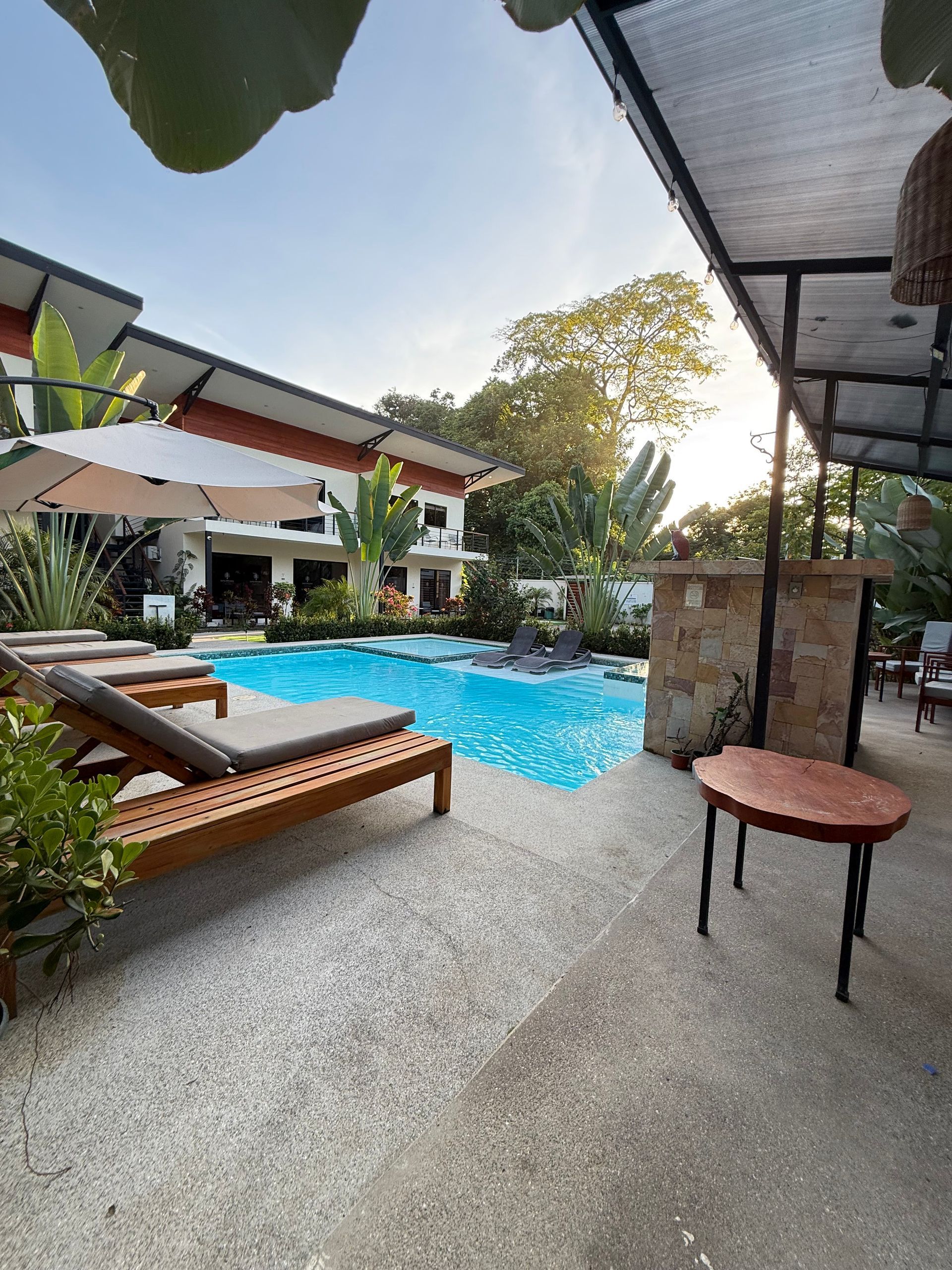 Pool area with lounge chairs, umbrella, bar, and two-story buildings in the background.