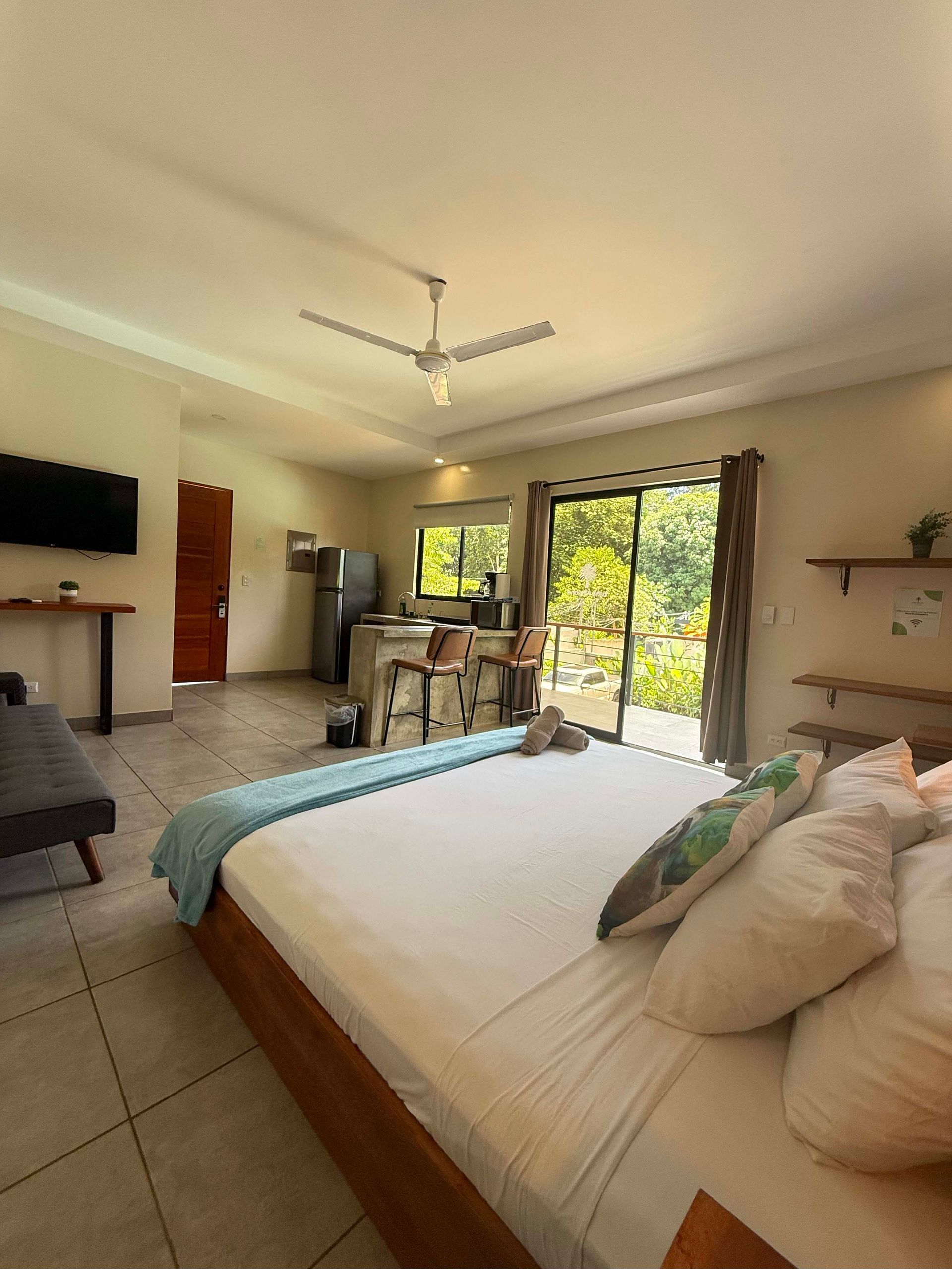 Bedroom with open layout, including bed, kitchenette, and balcony with trees.