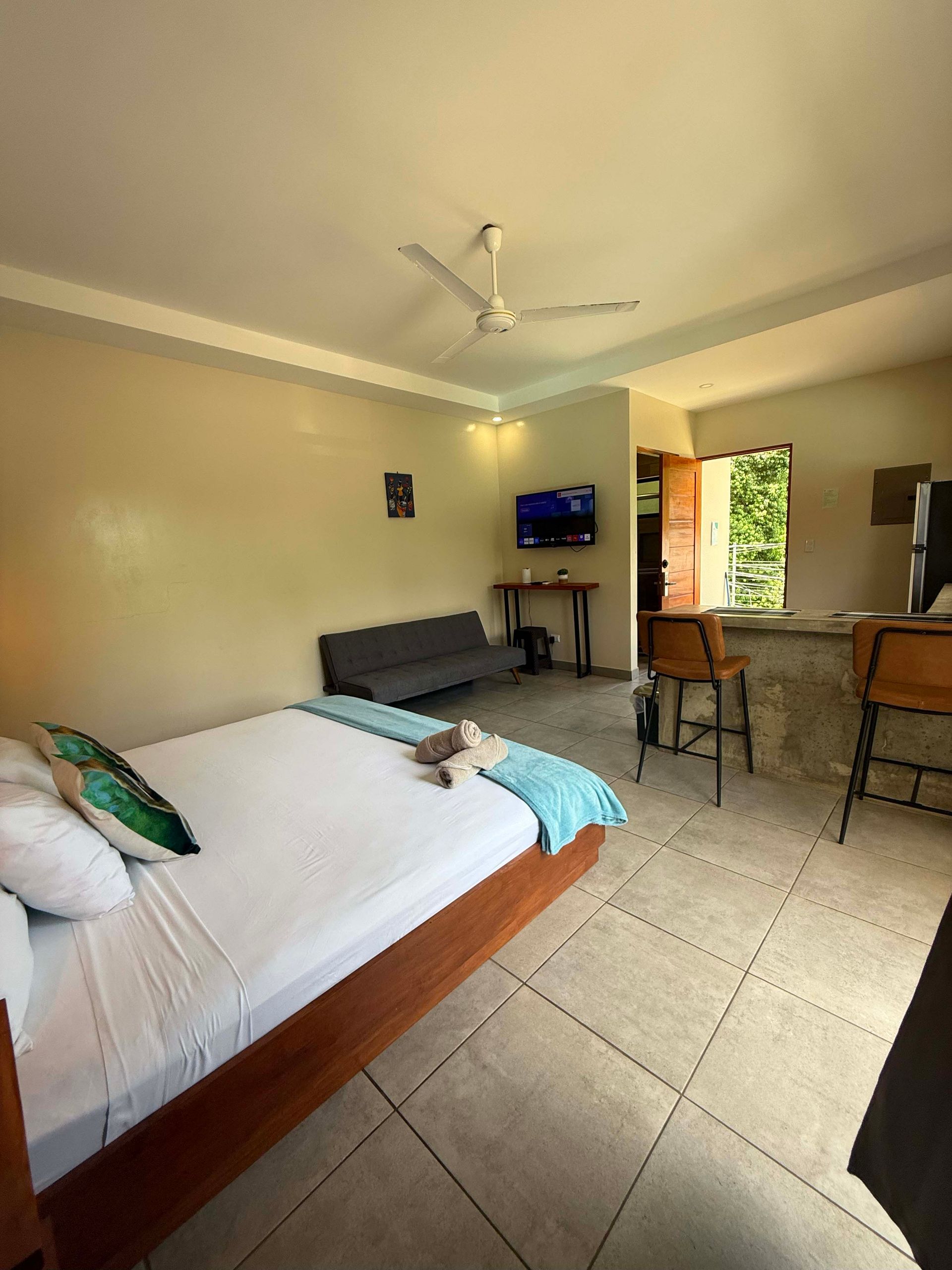 Cozy studio apartment with a bed, sofa, bar, and TV. Neutral walls and tiled floor.
