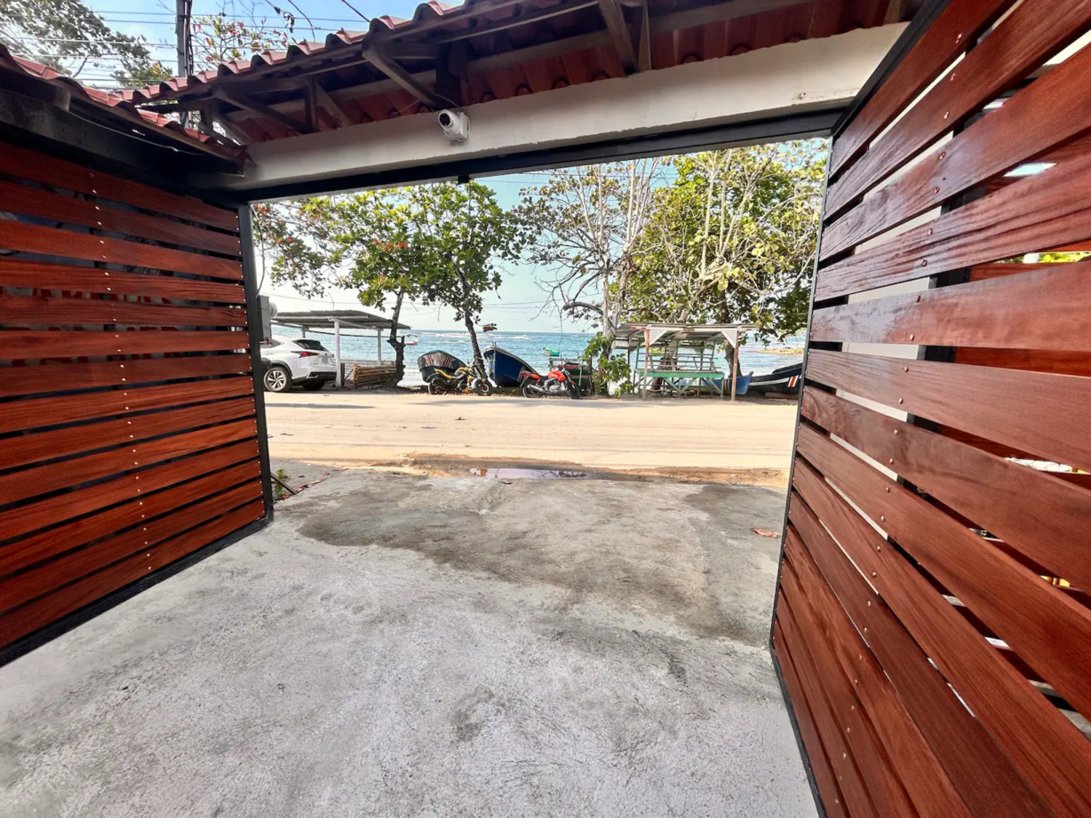 Open wooden gate framing a view of a street, beach, and parked vehicles.