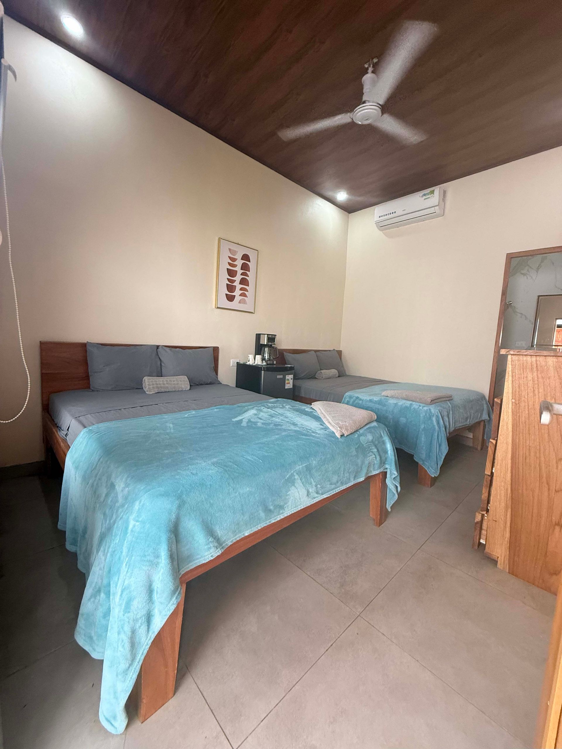 Bedroom with two beds, one larger and one single, with turquoise blankets. Beige walls, wood ceiling.