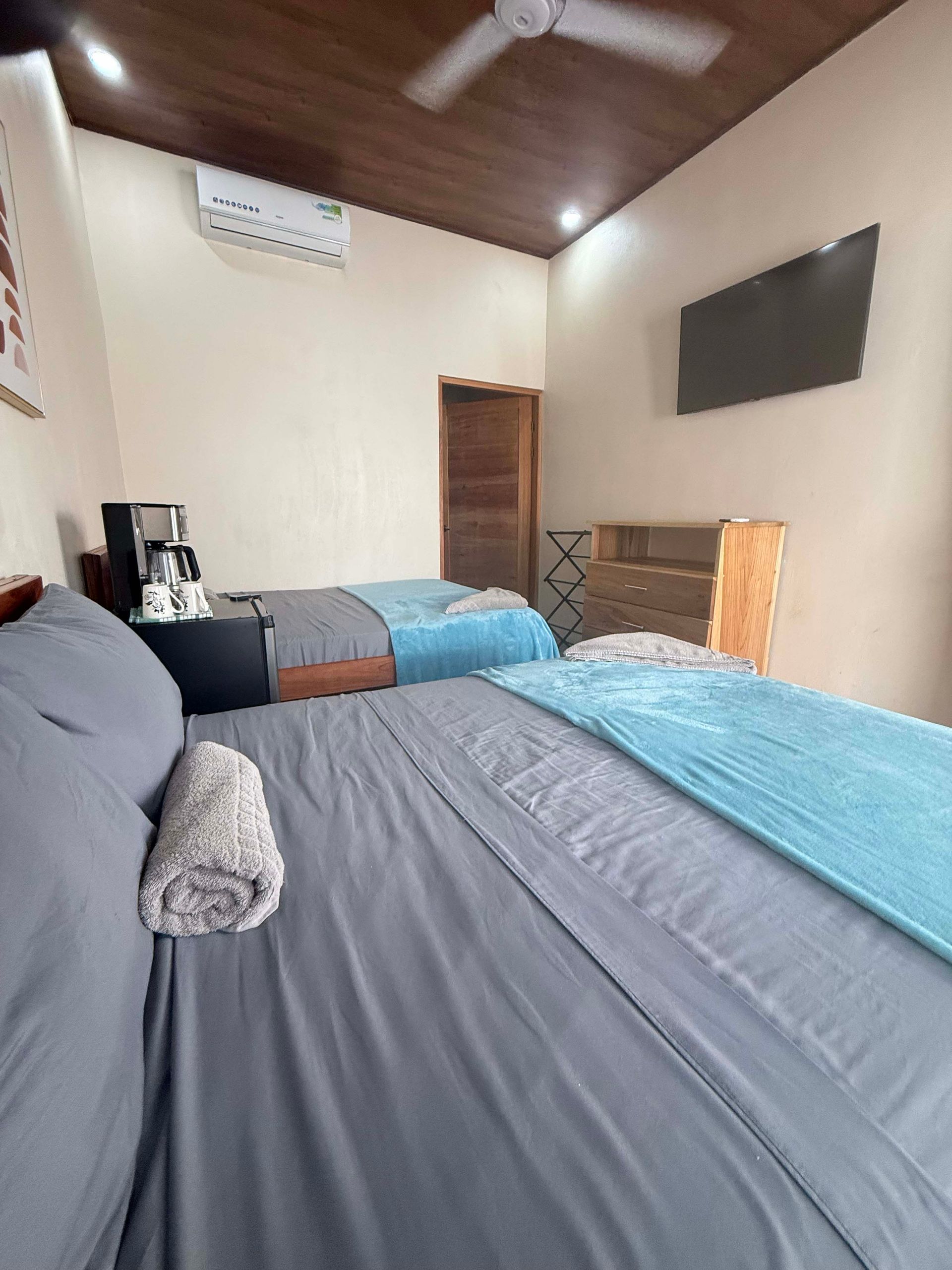 Bedroom with two beds, one gray, one blue; TV, air conditioner, coffee maker, and wooden ceiling.