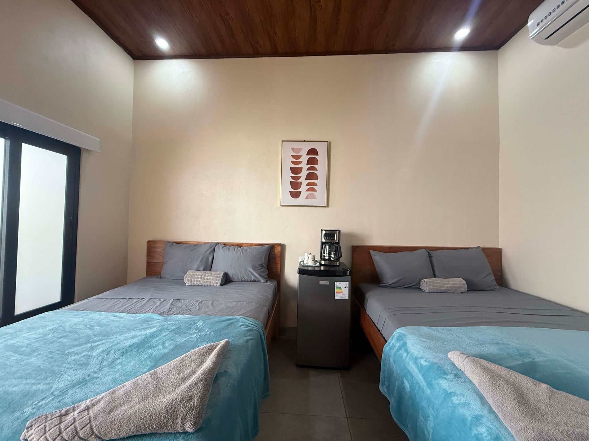 A hotel room with two beds, a mini-fridge, and a coffee maker. Turquoise and gray bedding. Wooden ceiling and frames.