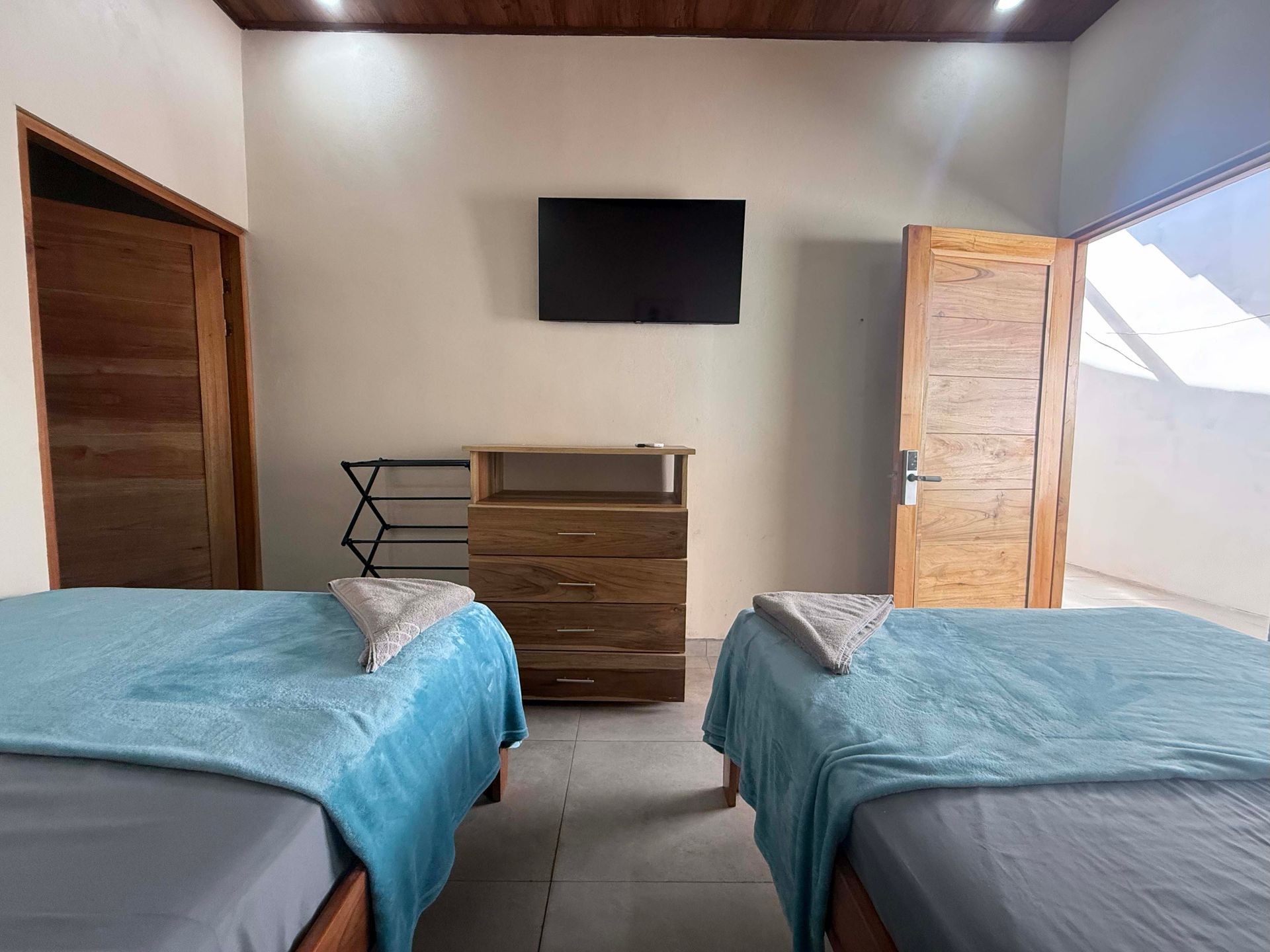 Two beds with teal blankets face a dresser with a TV above it, two doors on each side.