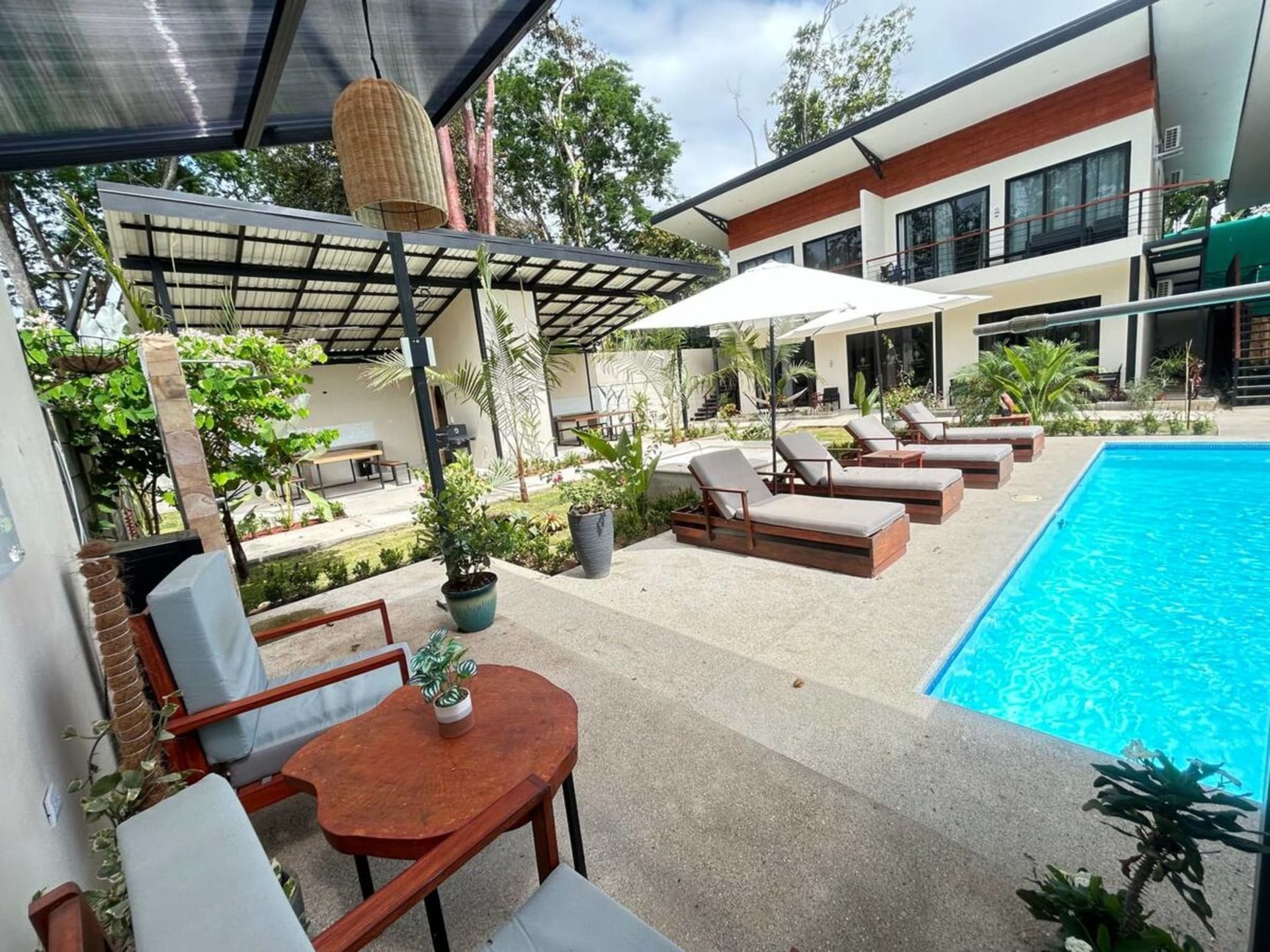 Poolside view: lounge chairs, umbrella, pool, modern house, patio furniture. Lush greenery.