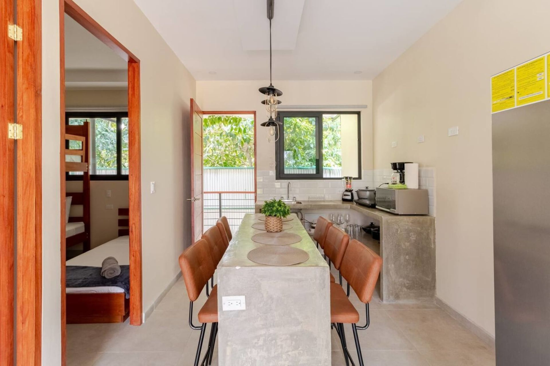 Dining area with concrete table, brown chairs, open door to balcony, and kitchen with appliances.