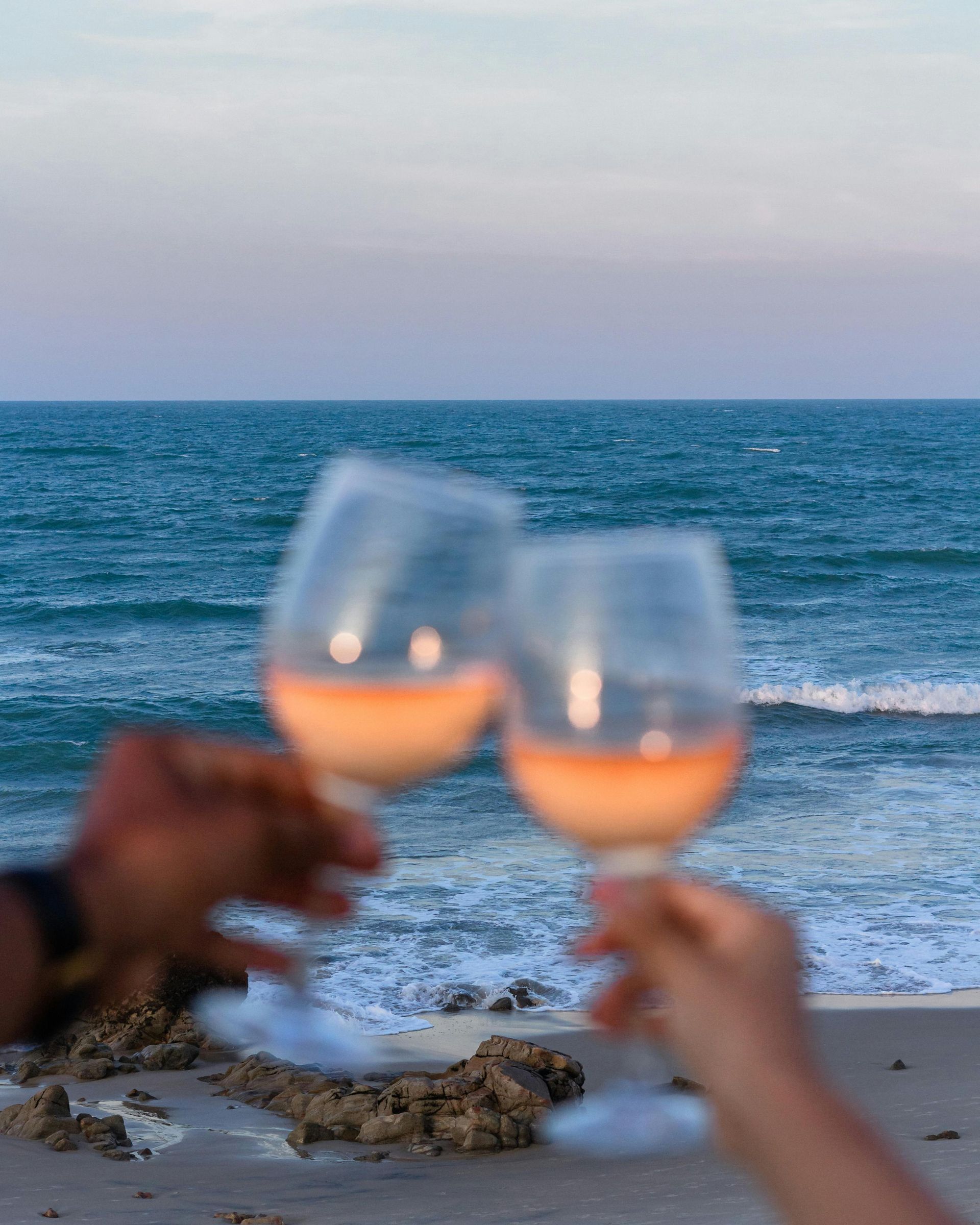 Two hands holding up wine glasses, toasting on a beach with ocean and sky in the background.