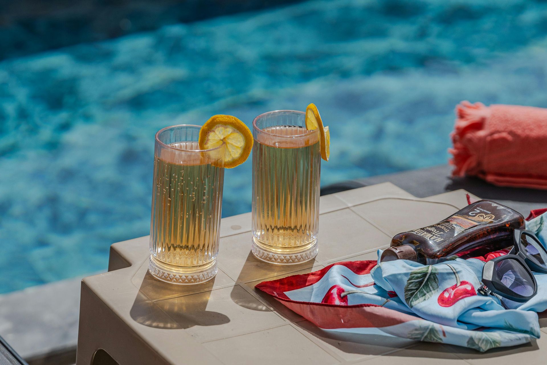 Two drinks with lemon slices next to a pool, sunglasses, and a towel.