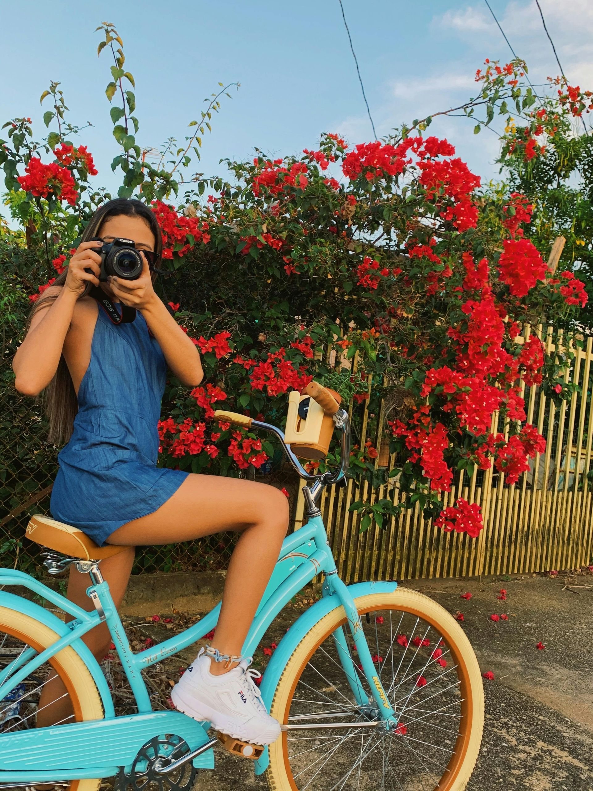 Woman in blue dress on blue bicycle, taking a photo in front of red flowers.