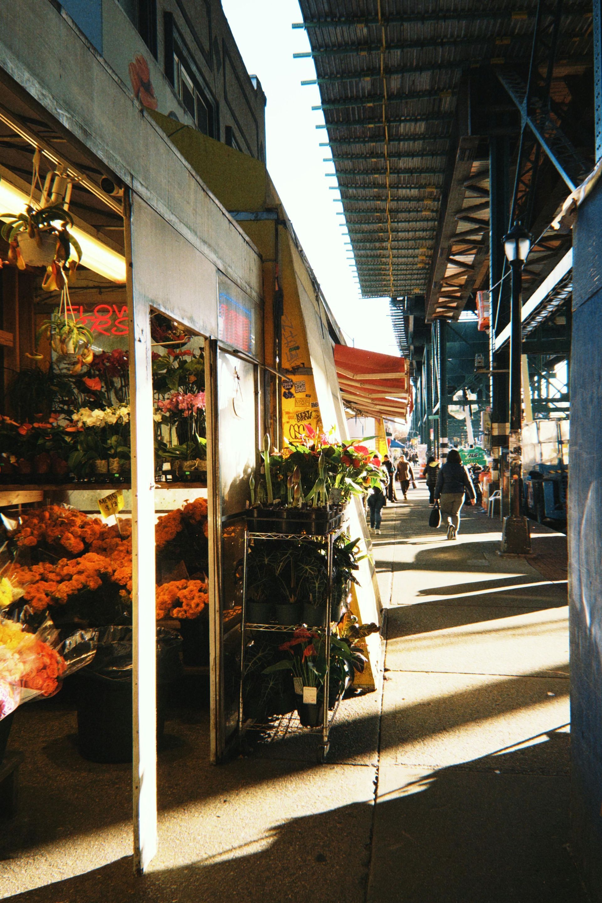 Flower stall with open doors under an elevated train track. Pedestrians walk in sunlight.