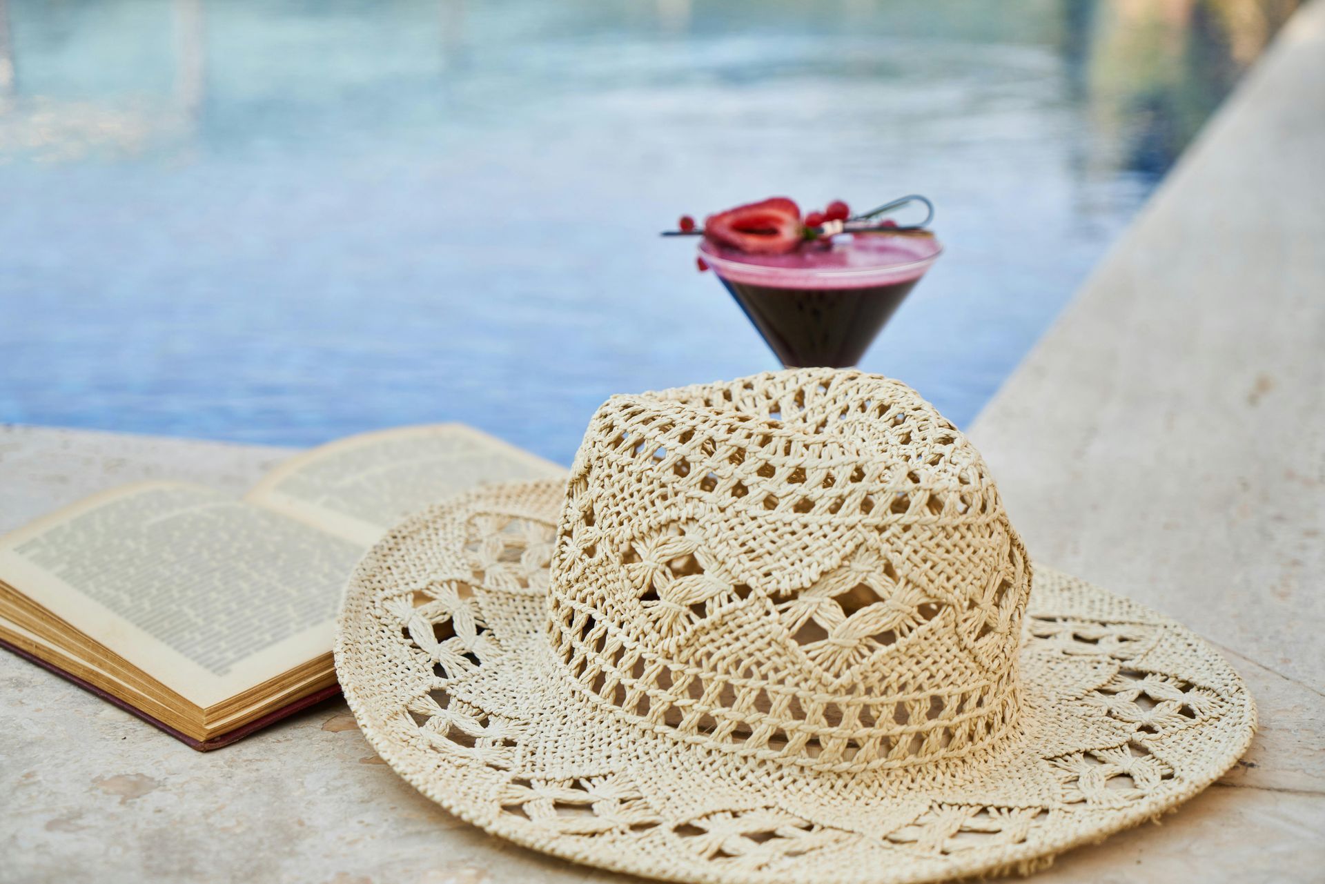 Straw hat, open book, cocktail with berries, poolside setting.