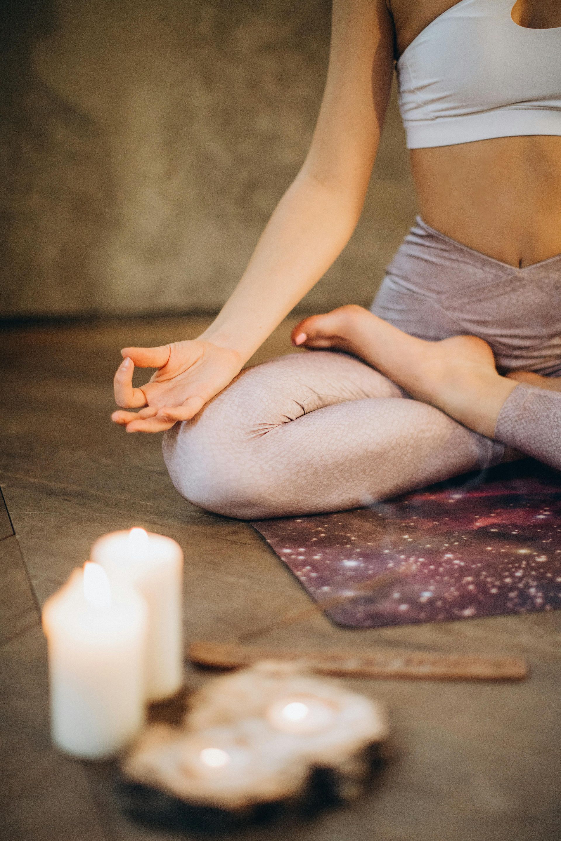 Person in yoga pose with hand gesture, sitting on a mat, candles in front.
