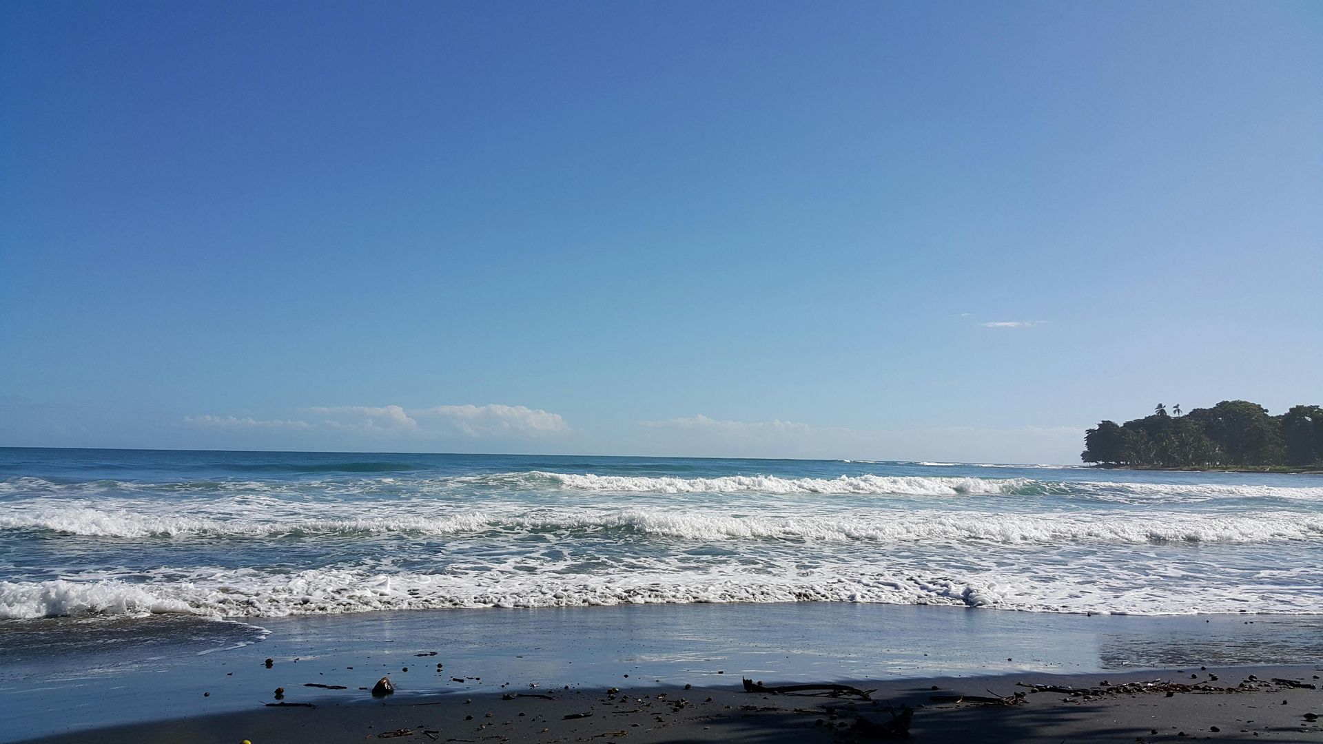 Ocean waves crashing on a dark sandy beach under a bright blue sky.