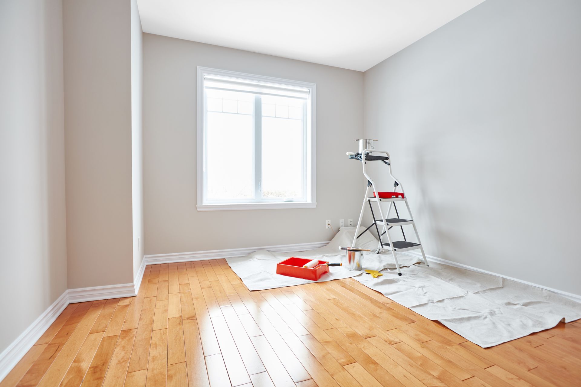 Empty room with hardwood floor, window, and painting supplies on drop cloths and ladder.