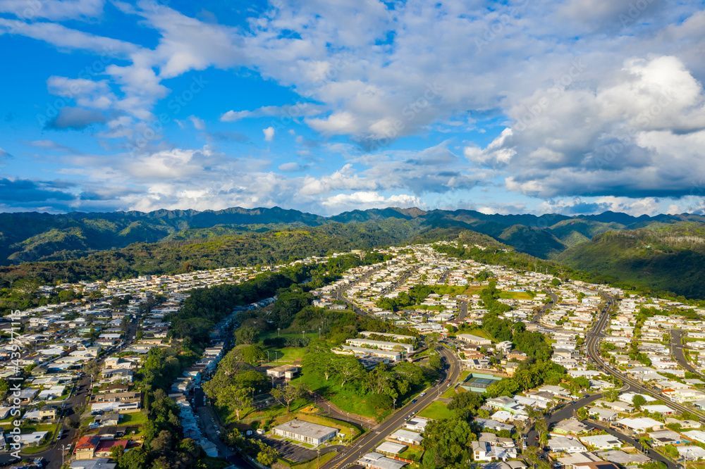 Aerial view of suburban houses nestled in a valley, with lush green hills in the background under a blue sky.