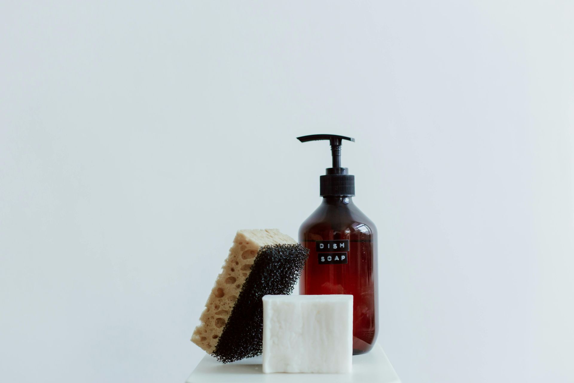 Brown soap dispenser, sponge, and white cotton pad on a white surface.
