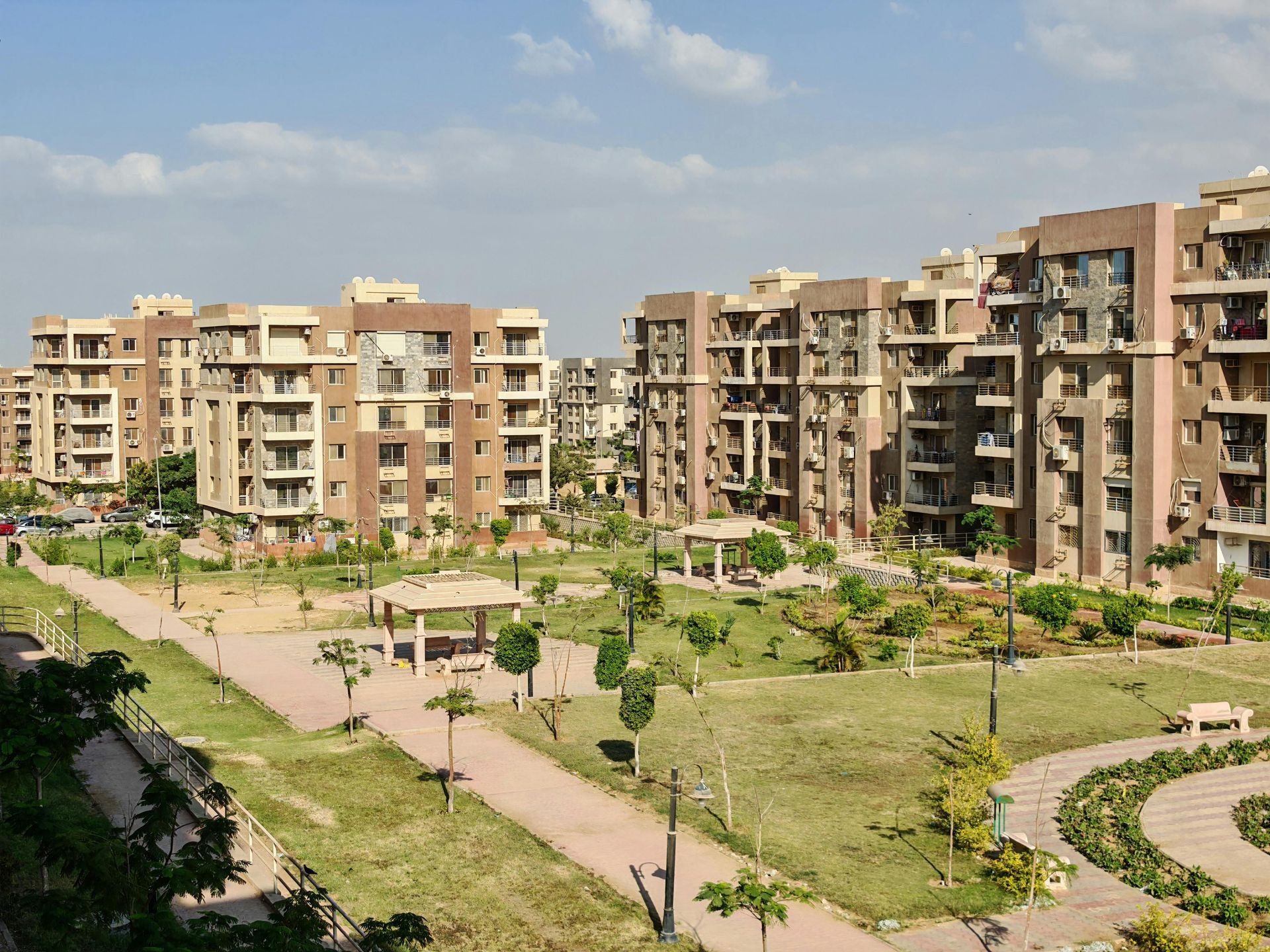 Apartment complex with multiple brown buildings, green lawn, walking paths, and a clear blue sky.