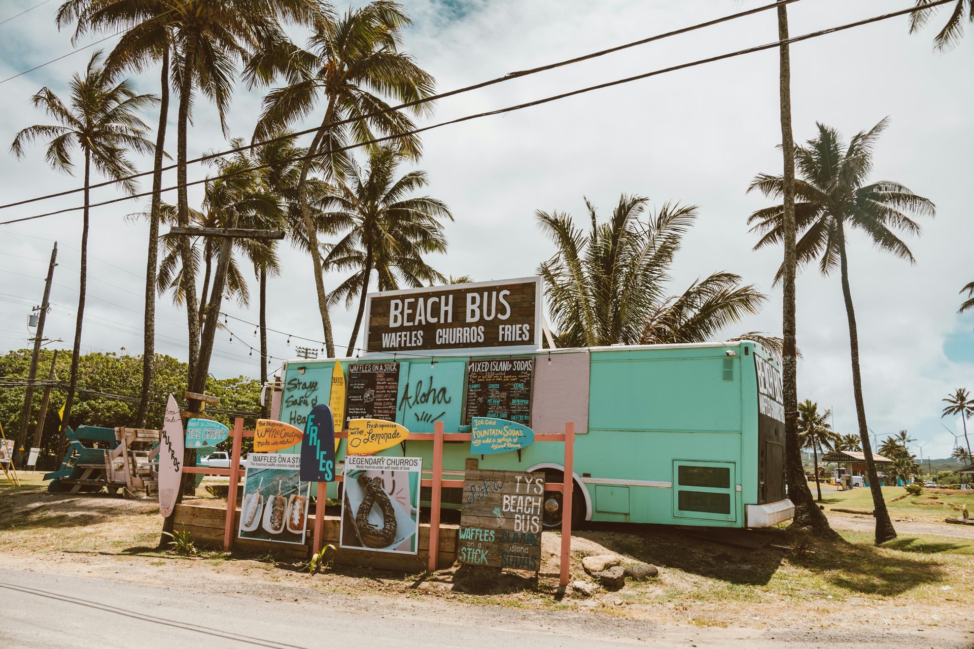 A teal beach bus with signs in front, palm trees overhead, on a road.