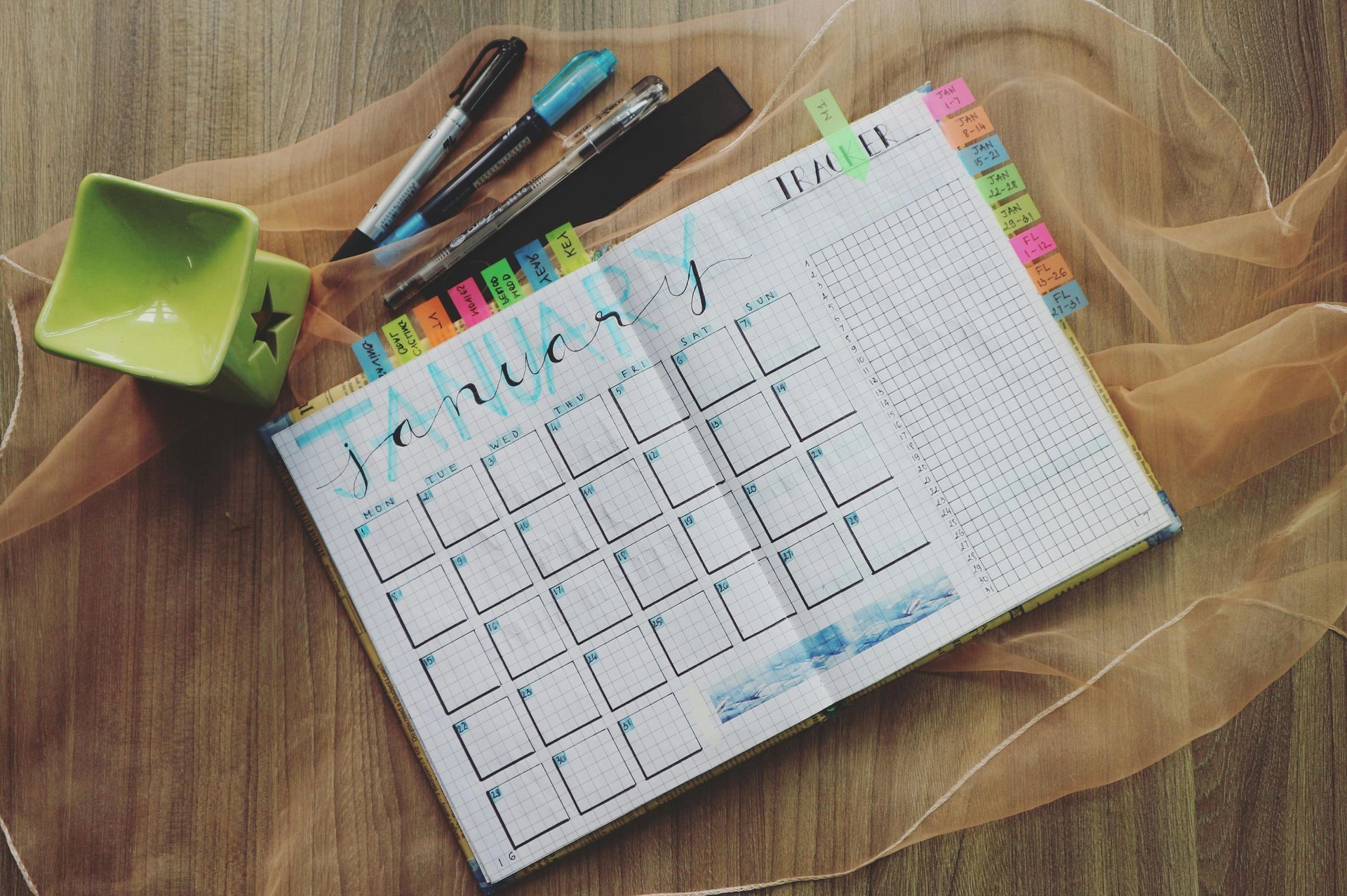 Calendar open to January, with colorful tabs, pens, and a decorative cup, on a wooden surface.