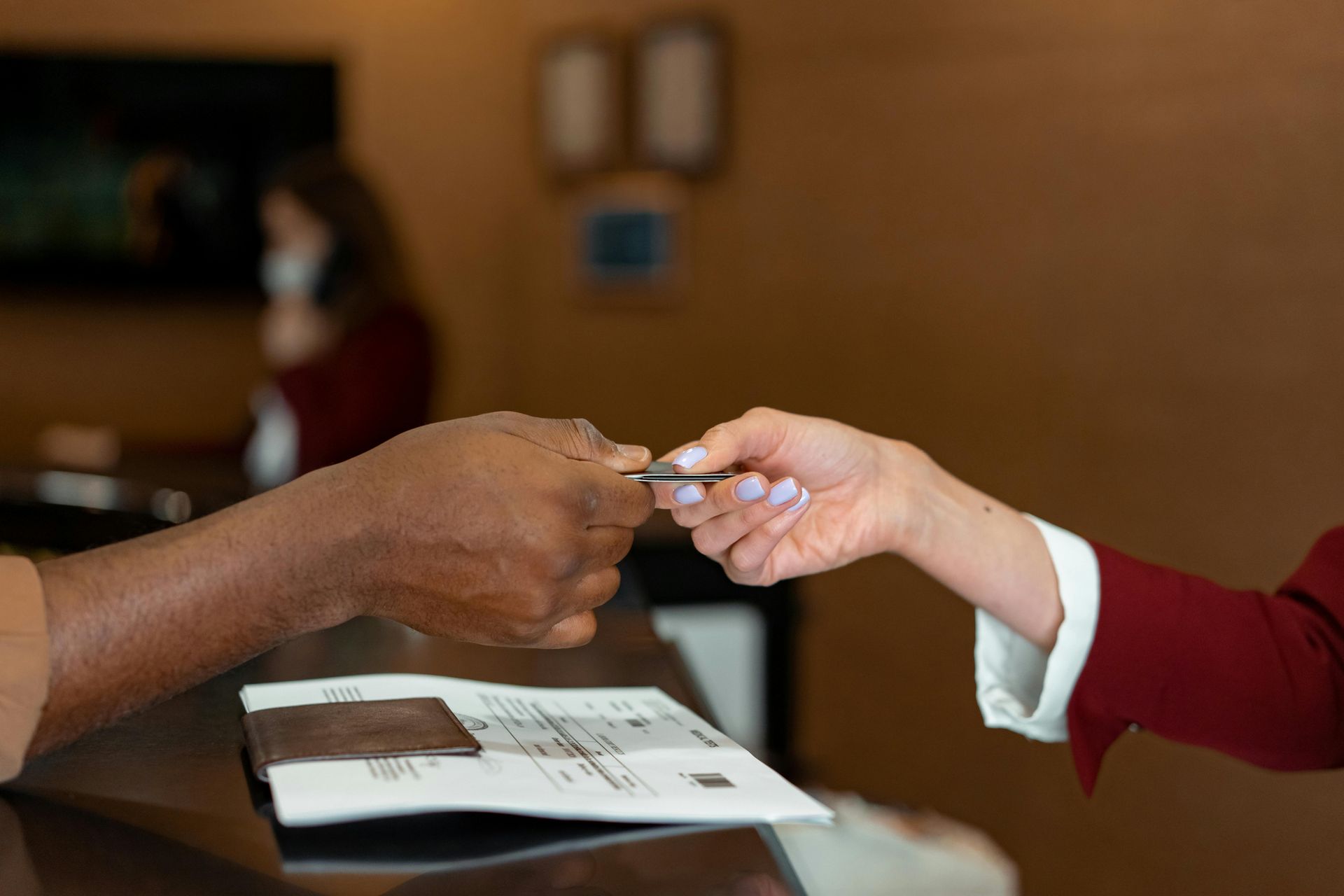 Hands exchanging a key card over a hotel reception desk. A person wearing a mask is in the background.