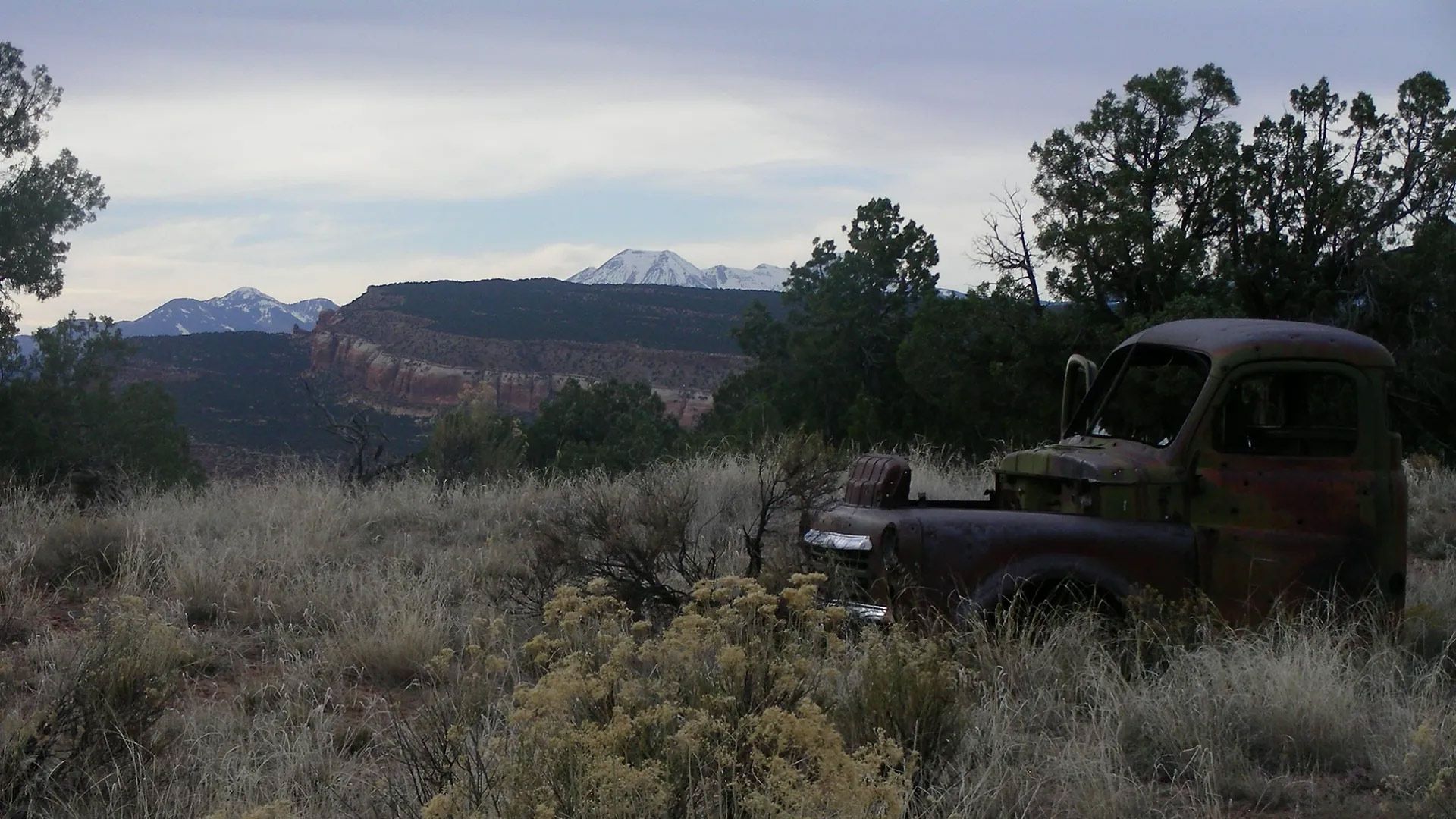 An old rusty truck is parked in a field with mountains in the background.