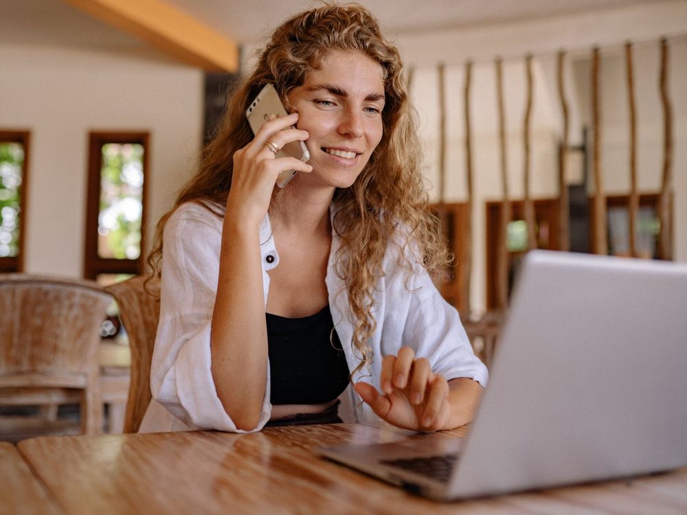 A woman is sitting at a table talking on a cell phone while using a laptop computer.