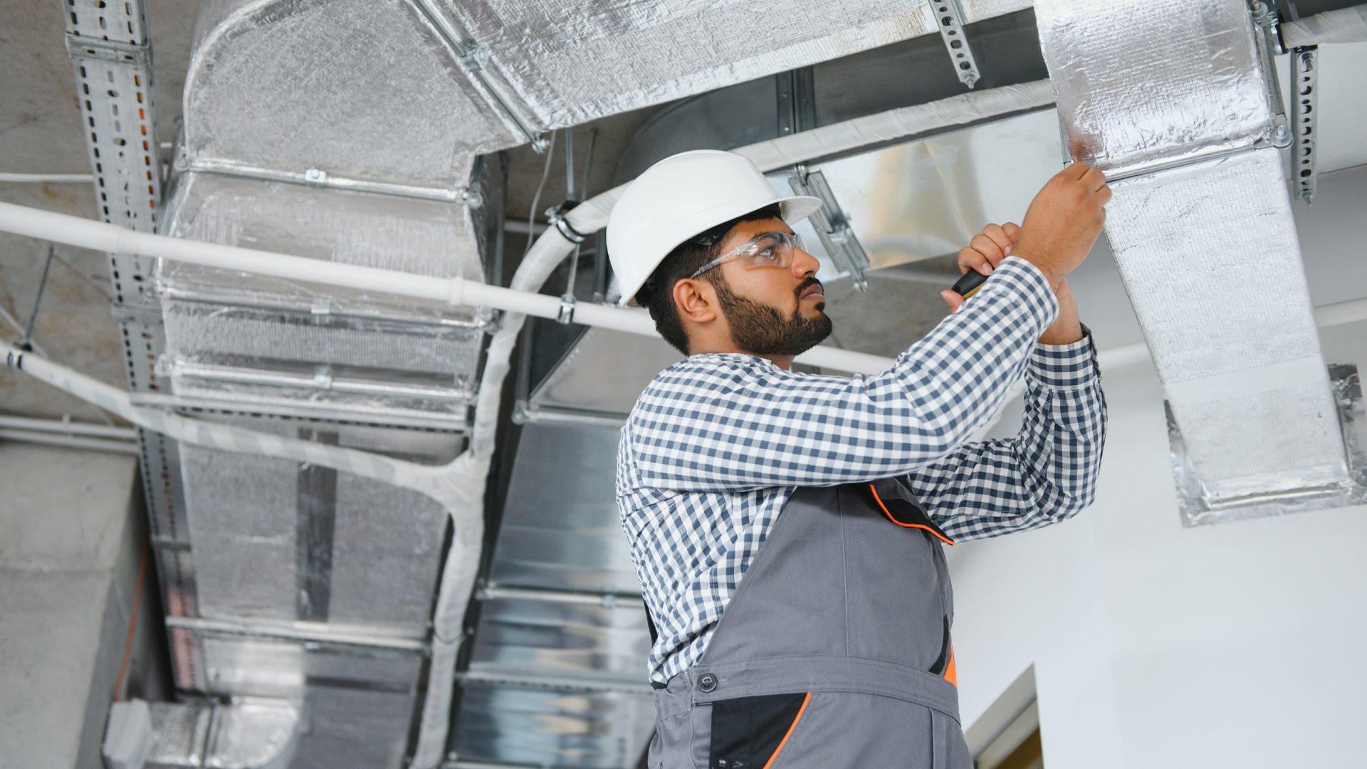 HVAC worker in hard hat inspecting ductwork; interior setting.