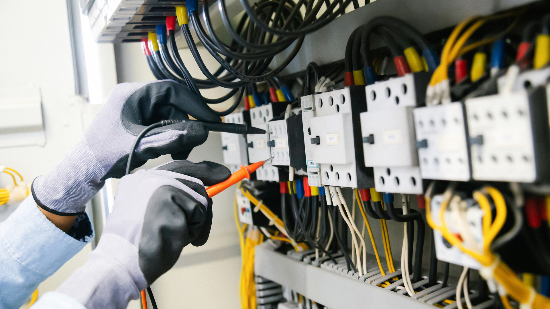 Electrician testing electrical panel wires with a multimeter, wearing gloves.