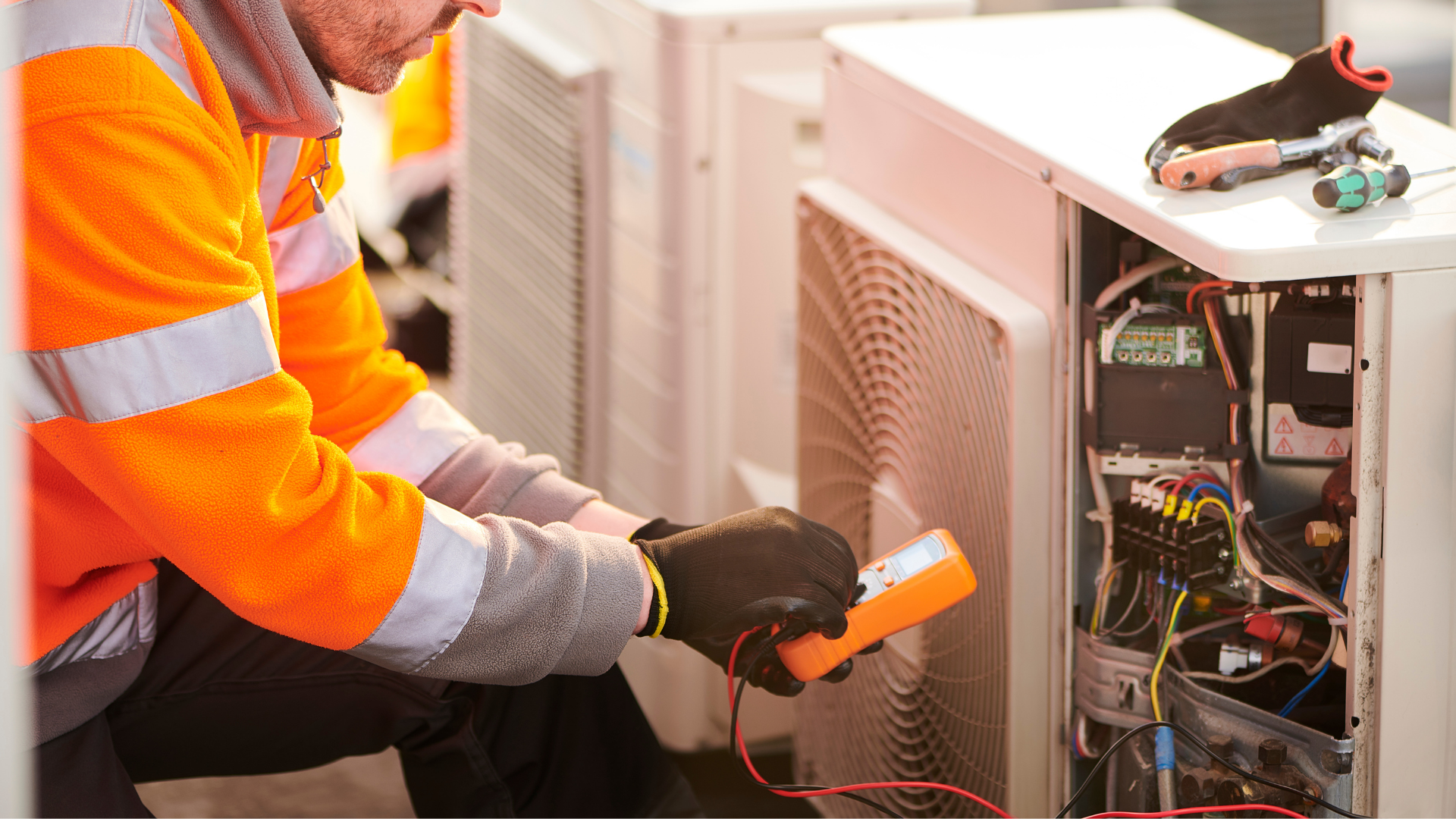 HVAC technician in an orange safety vest, using a multimeter on an outdoor unit.