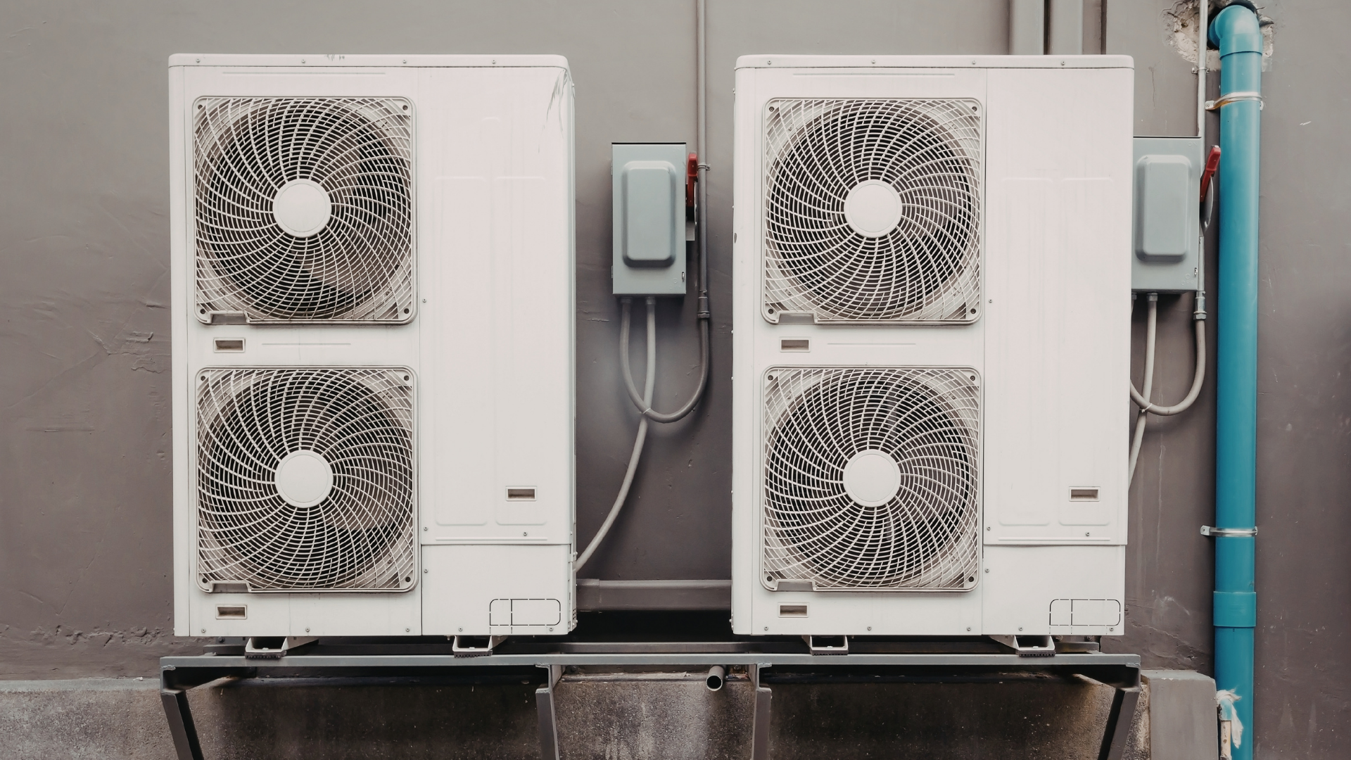 Two white HVAC units with fans, mounted on a gray wall, blue pipes, and electrical boxes.