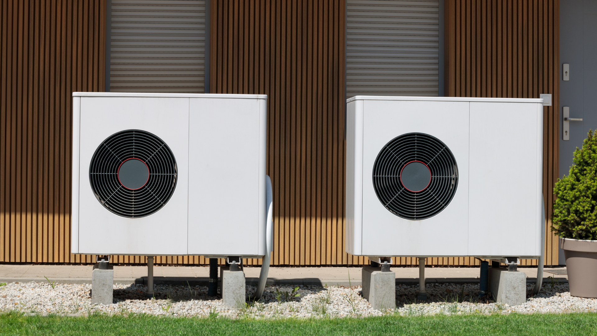 Two white heat pump units on concrete blocks in front of a wooden-paneled building.