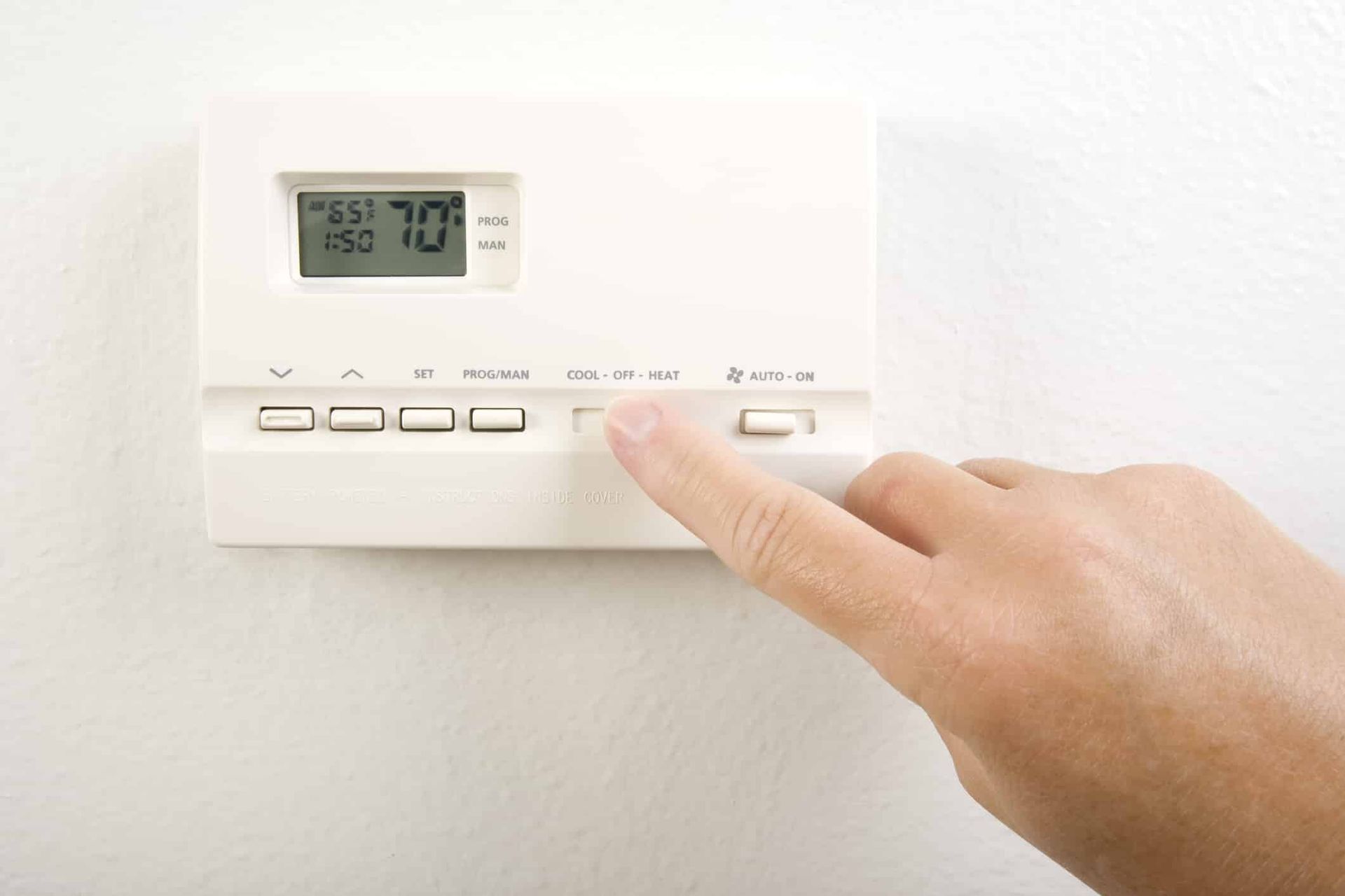 Hand adjusting a white thermostat on a white wall; display shows 70 degrees.
