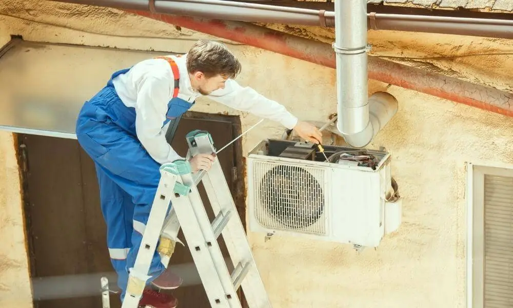 Man in blue overalls on a ladder repairs an outdoor air conditioner unit on a building.