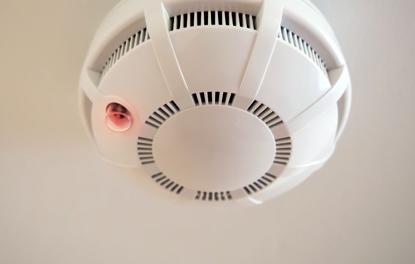 White smoke detector mounted on a white ceiling, with a red light visible.