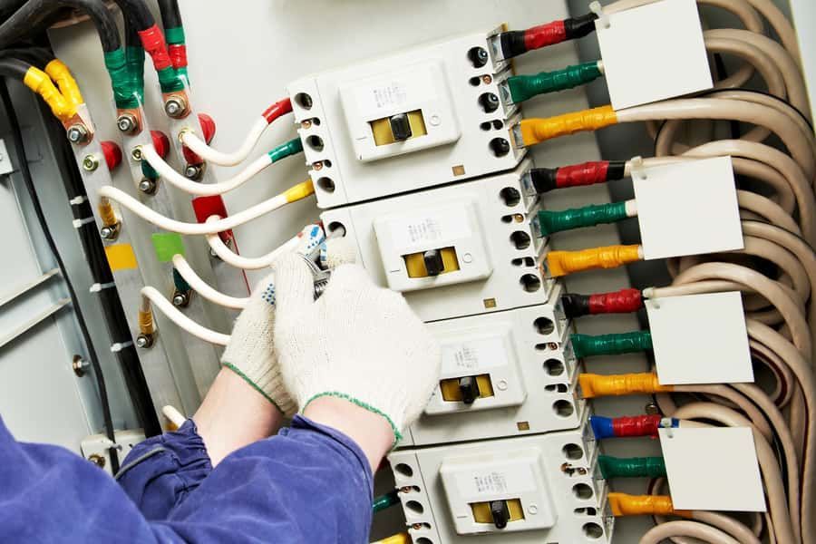 Hands in work gloves working on electrical wiring in a panel, various colored wires visible.