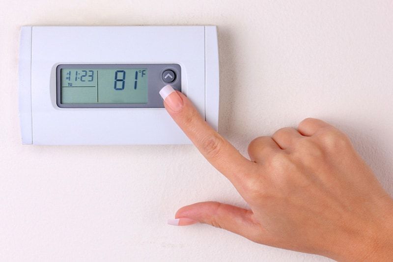 Hand pressing up arrow on a thermostat set to 81°F, mounted on a white wall.