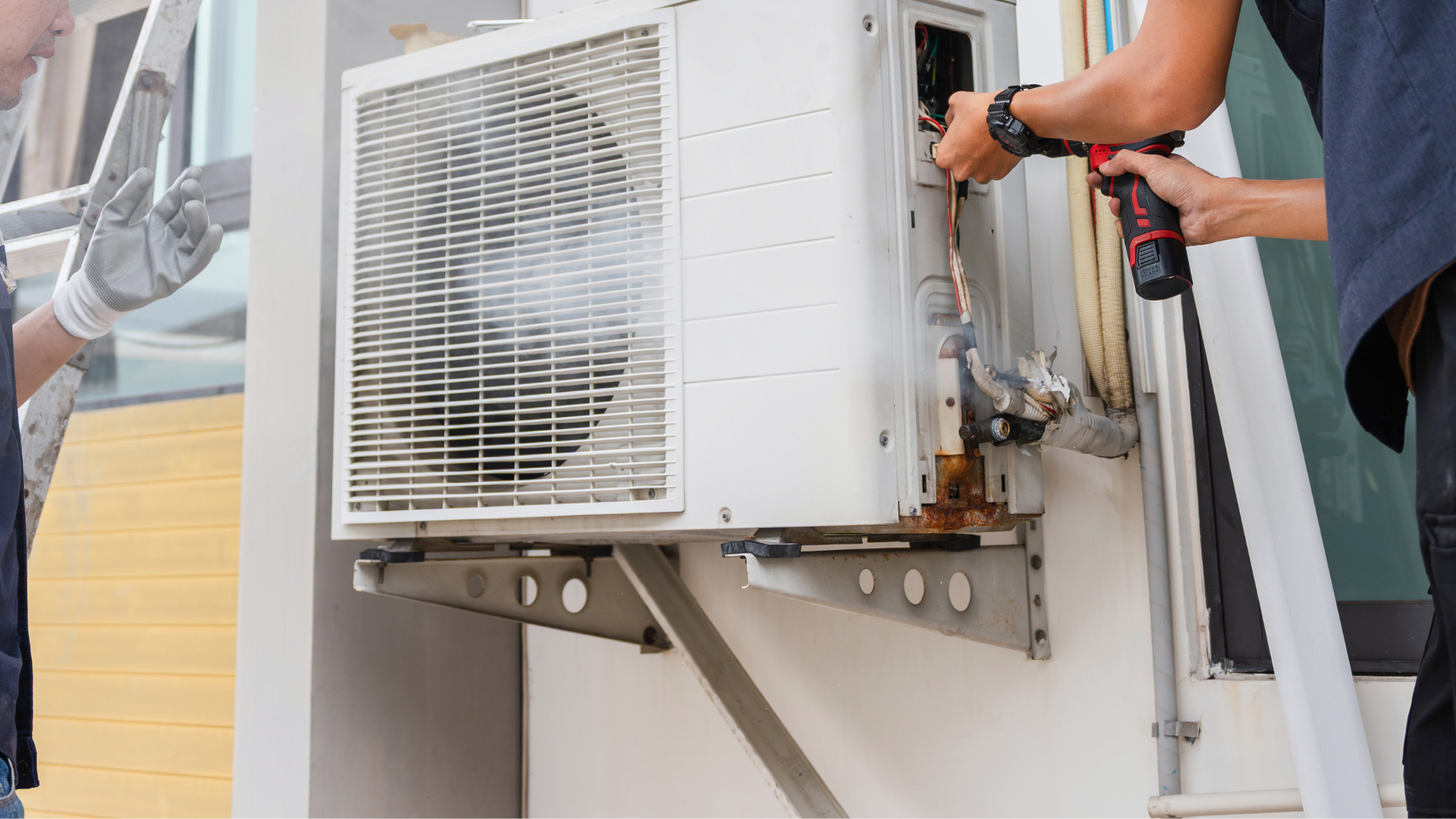 Two workers repairing an outdoor air conditioning unit attached to a building's wall.