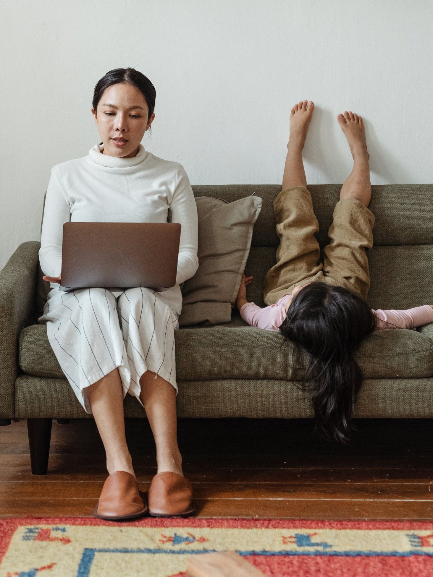 A woman is sitting on a couch using a laptop and a child is laying on the couch.