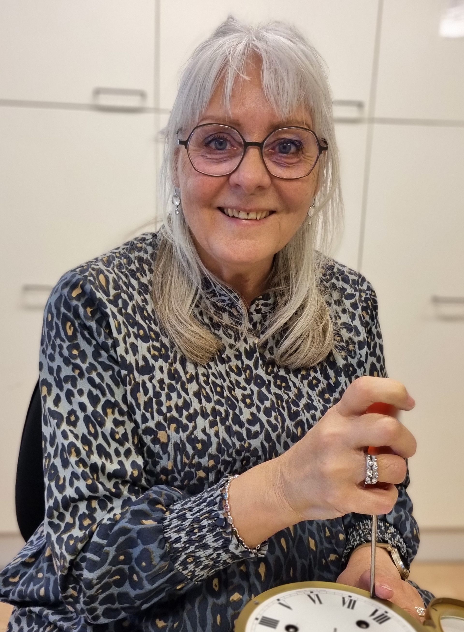 Kvinde med briller smiler ved siden af ​​en hængende køkkenvægt, mens hun holder en rød frugt i et lyst køkken.